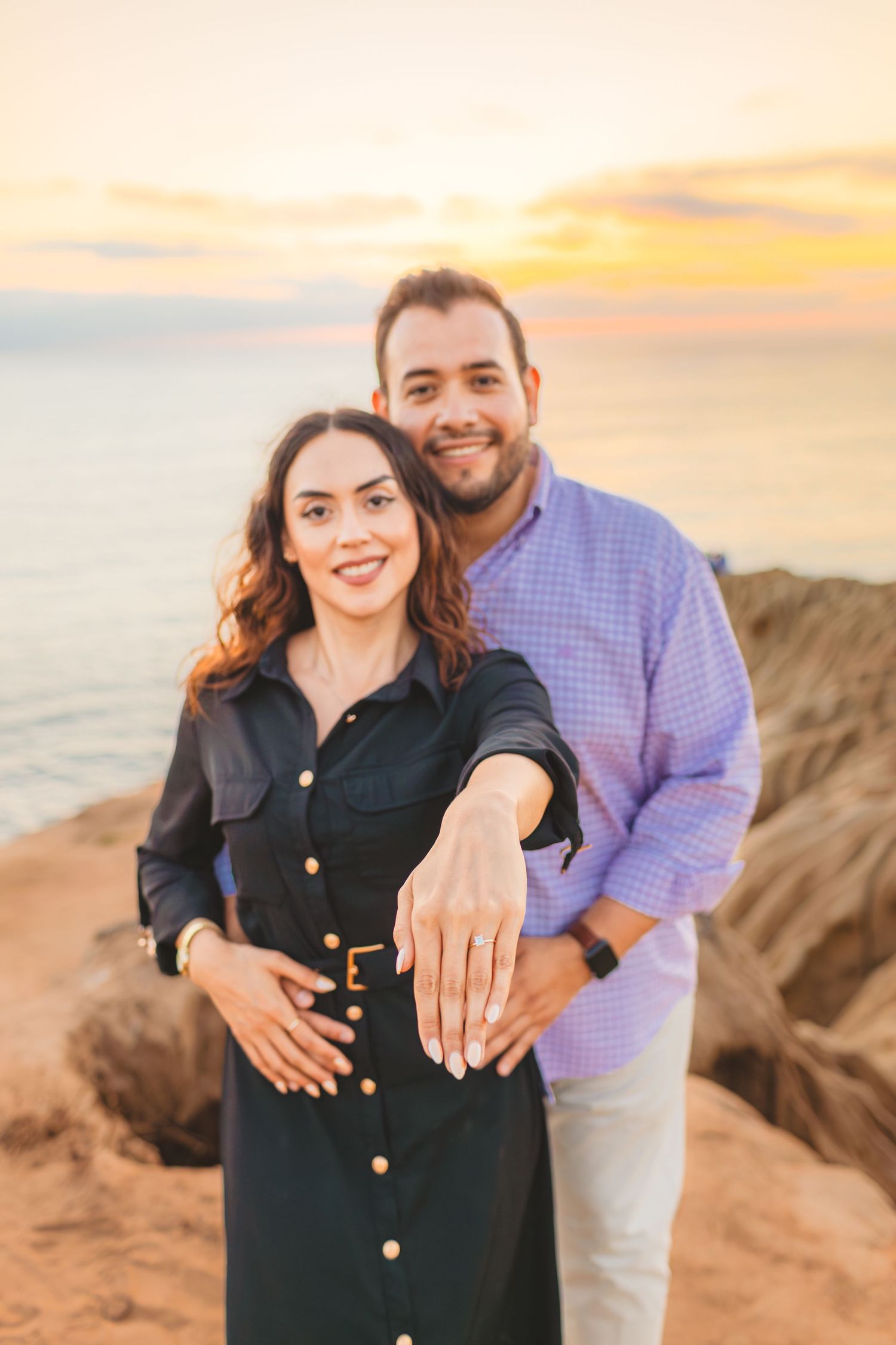 A couple poses together on a beach cliff at sunset, showing off an engagement ring.