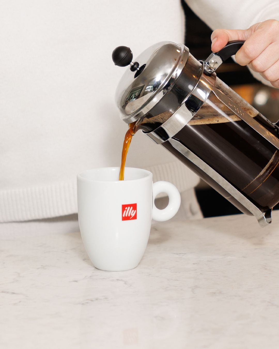 Coffee being poured from French press into white illy cup on marble counter.