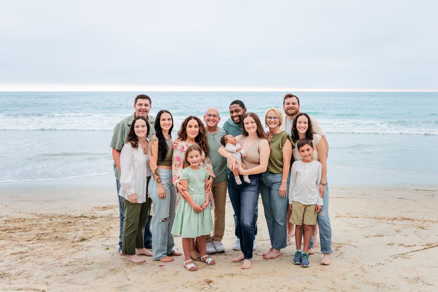 Large family gathering on sandy beach with ocean waves and cloudy sky in background.