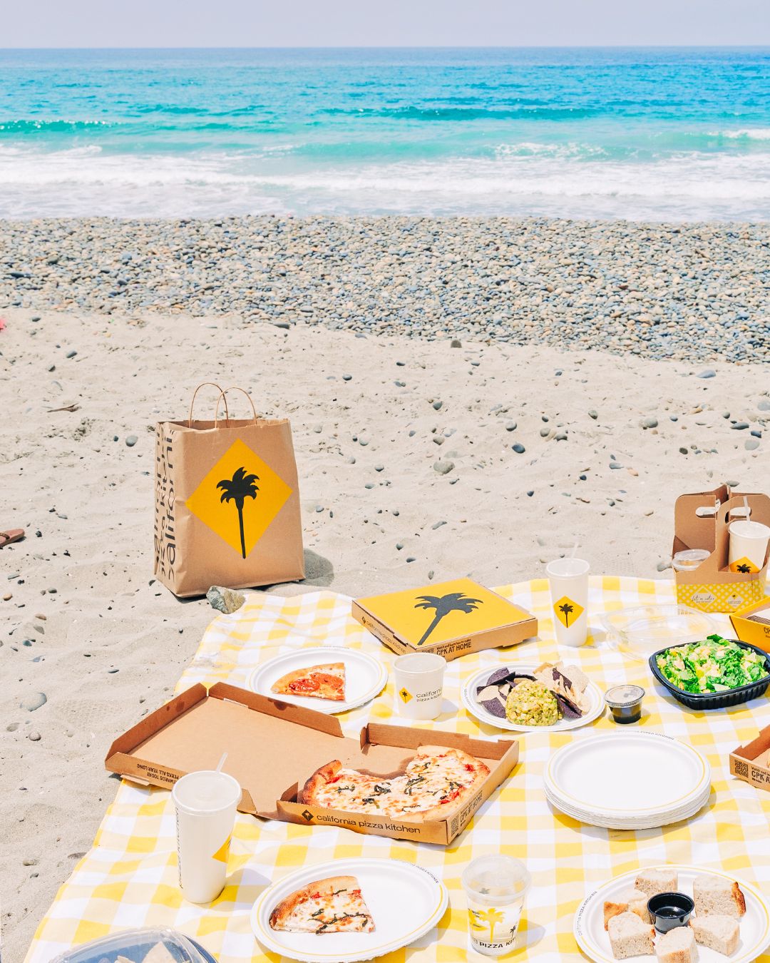 Beach picnic spread with pizza boxes and dishes laid out on yellow gingham blanket by the ocean.