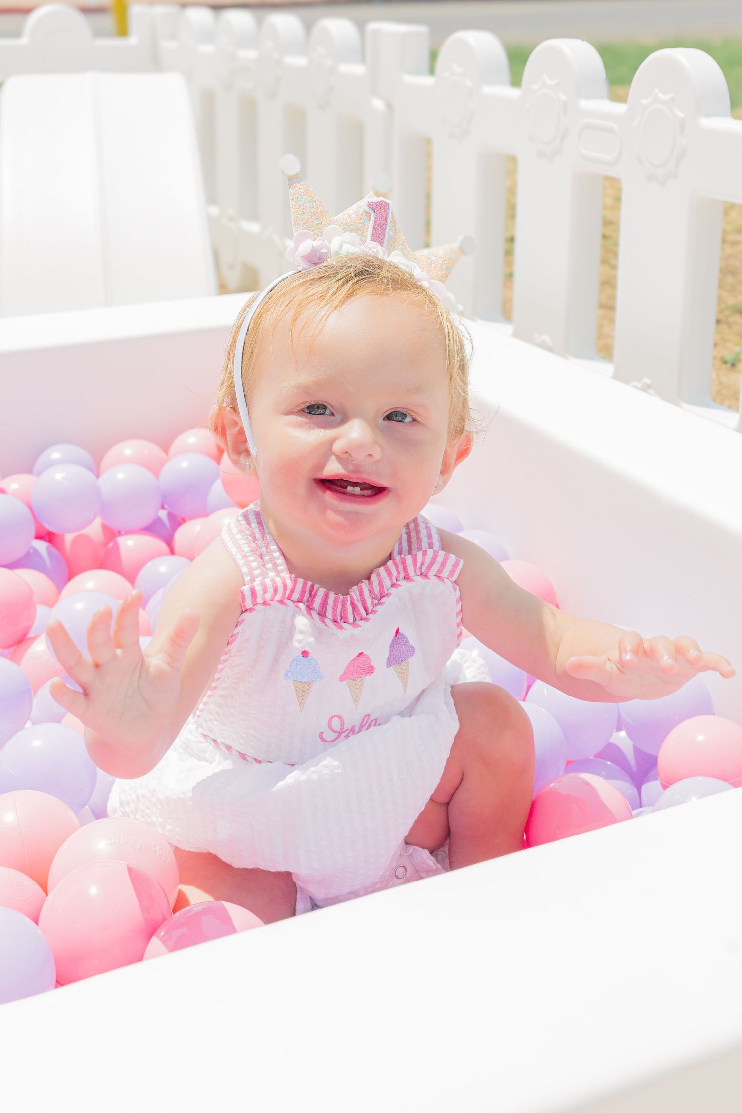 Smiling baby sitting in white bathtub filled with pink and purple balls during outdoor photoshoot.