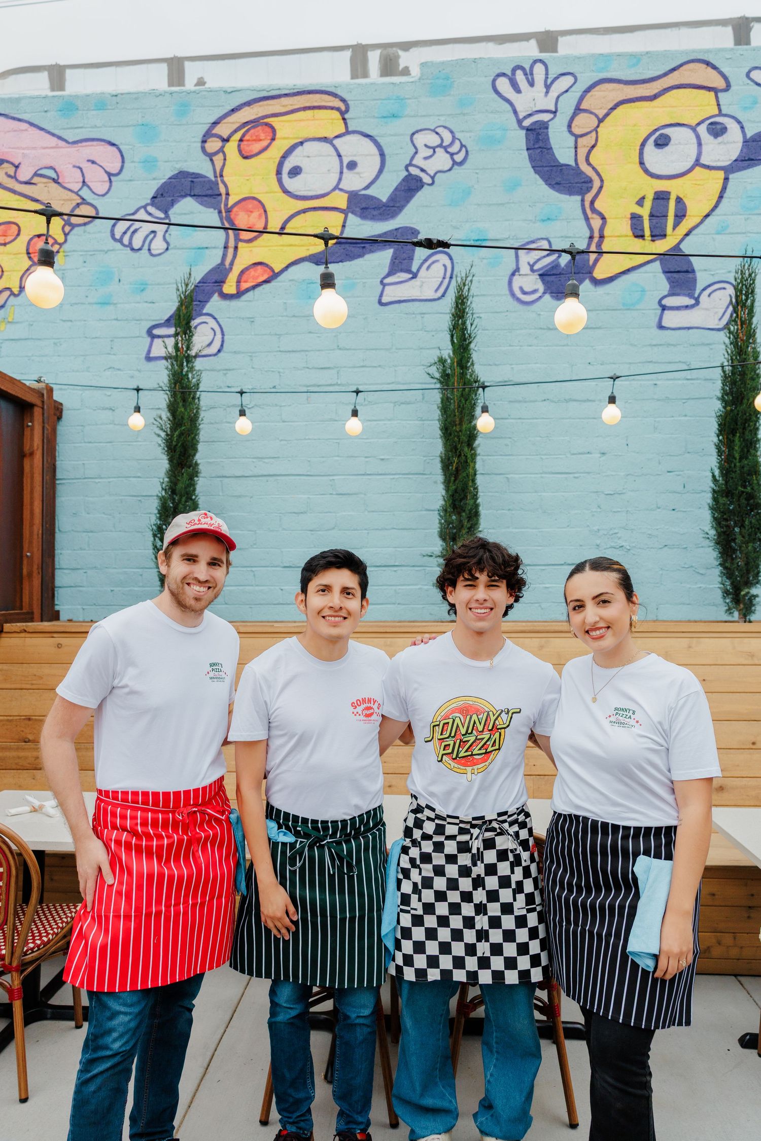 Restaurant staff wearing white shirts and aprons pose in front of a colorful mural wall with string lights.