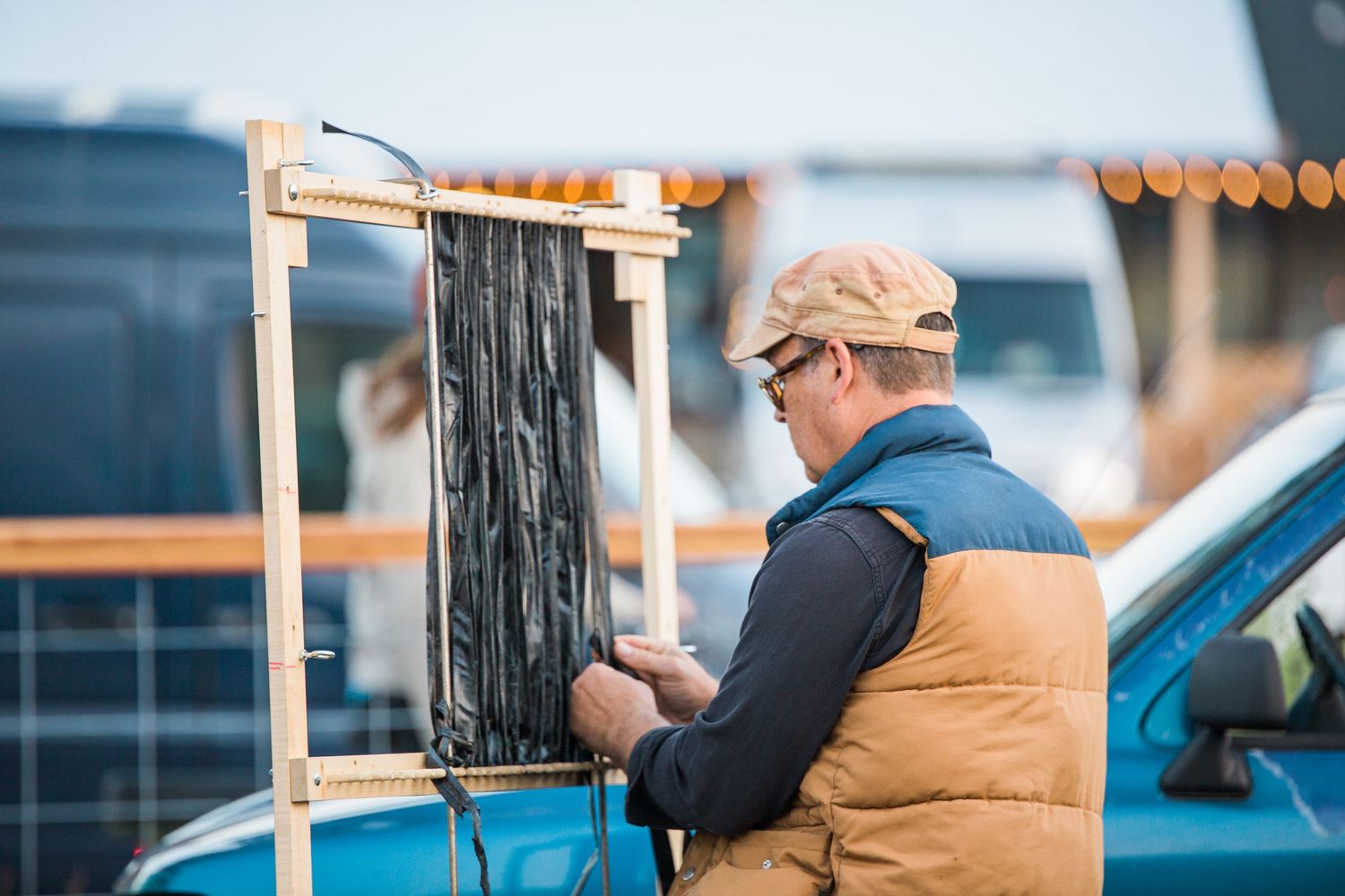An elderly person in a tan coat stands near railings at a pier with fishing equipment in natural lighting.