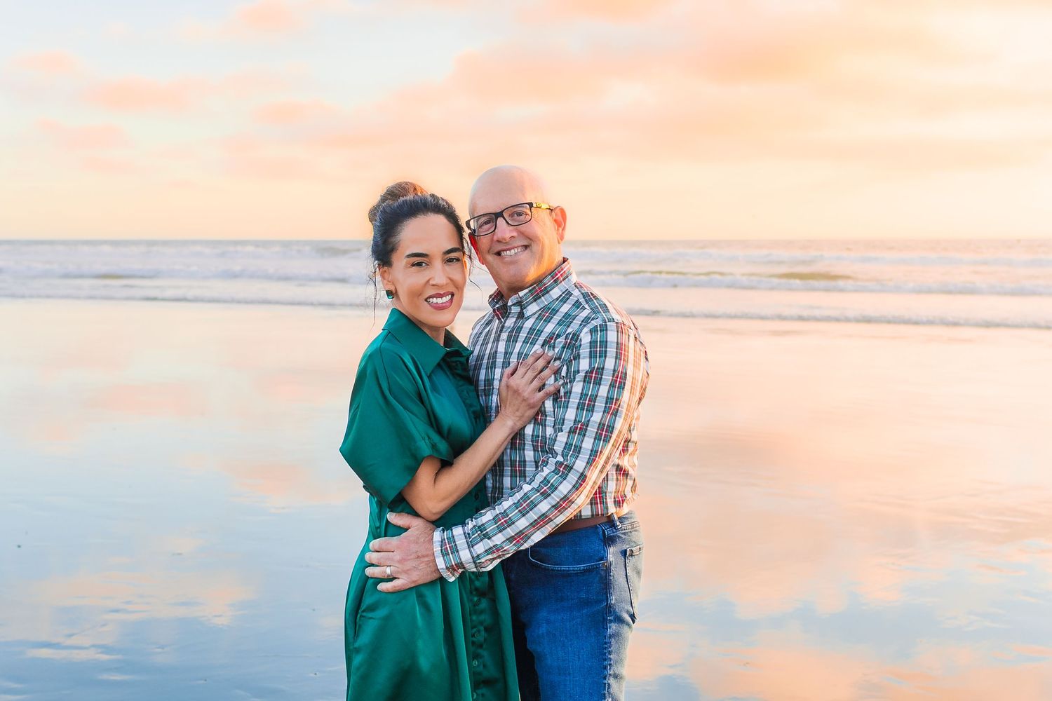 A couple embraces on the beach at sunset with soft pink and orange sky reflecting on the ocean water.