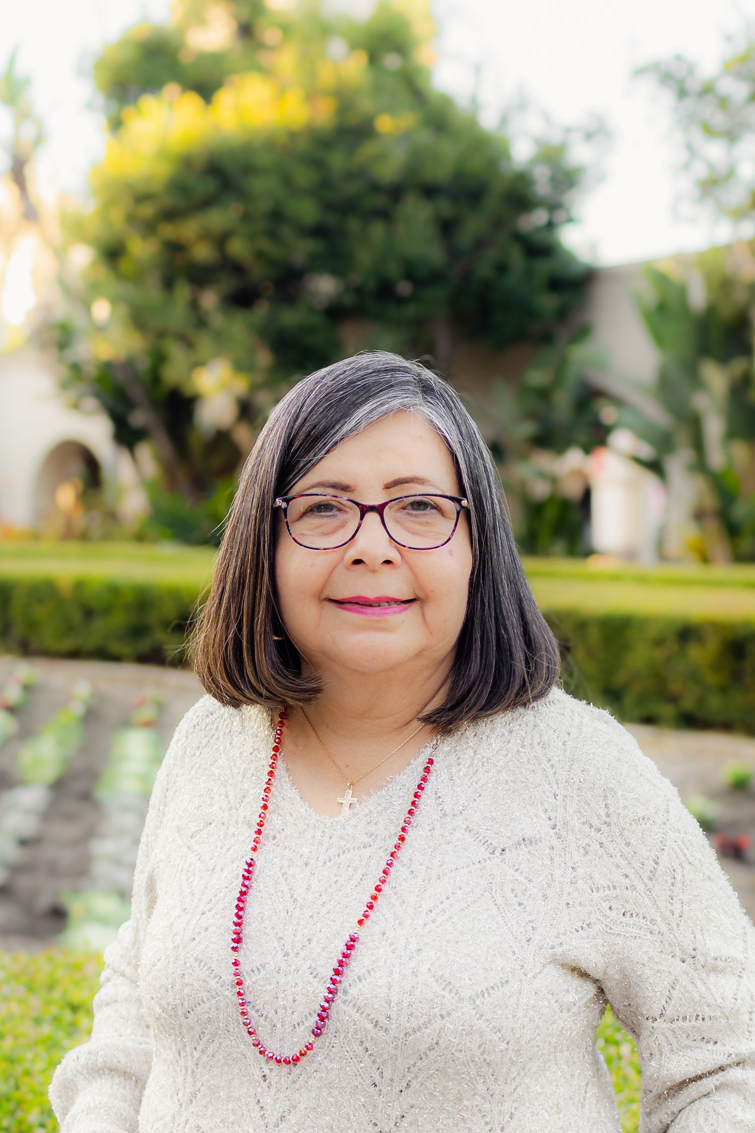 A person wearing glasses and a cream sweater with a pink beaded necklace poses in an outdoor garden setting.