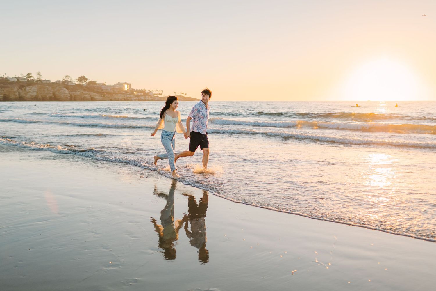 People walking along shoreline at sunset with waves crashing and warm orange sky.