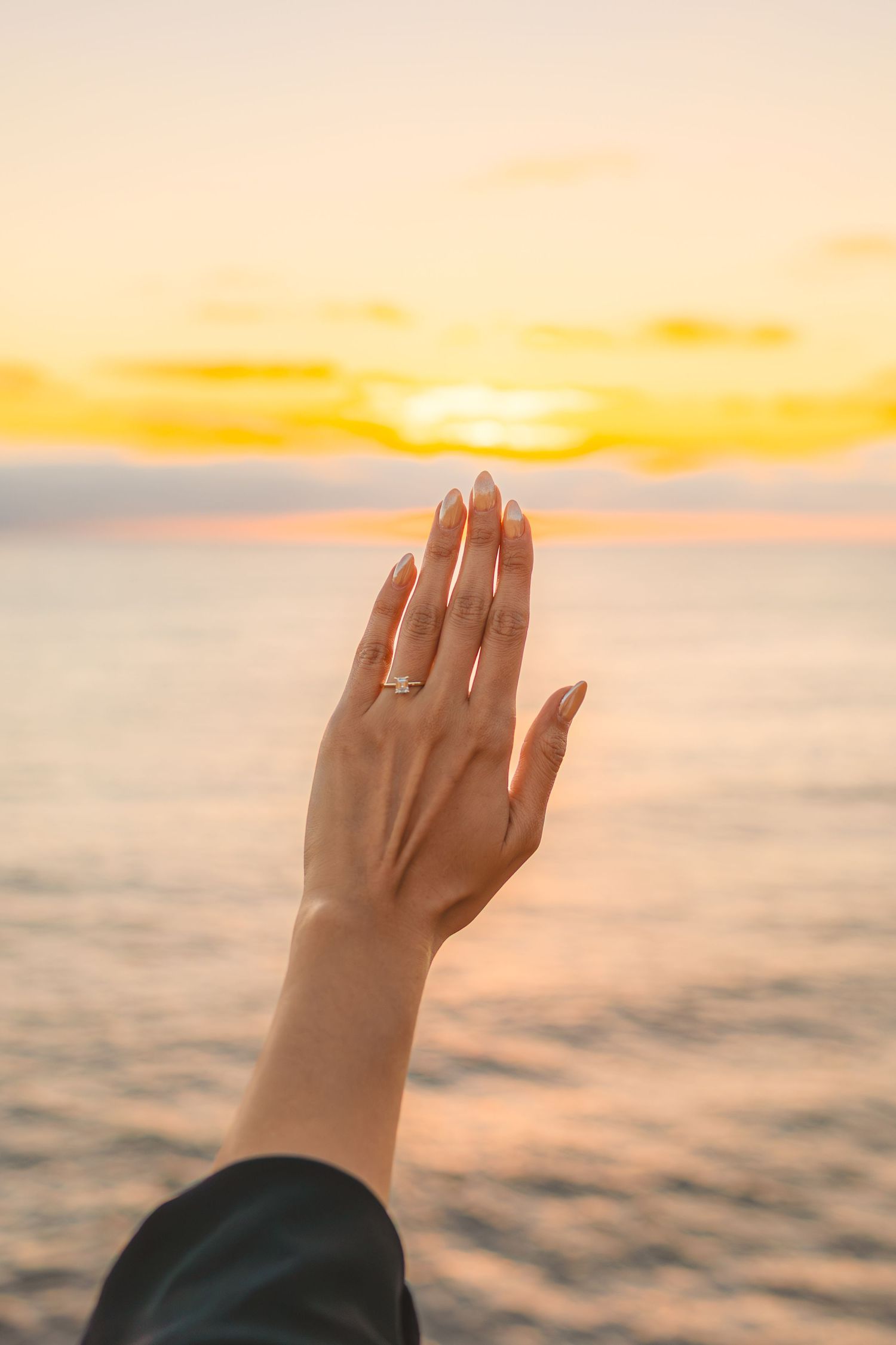 A hand with a ring reaches up towards a golden sunset sky over calm ocean waters during dusk.