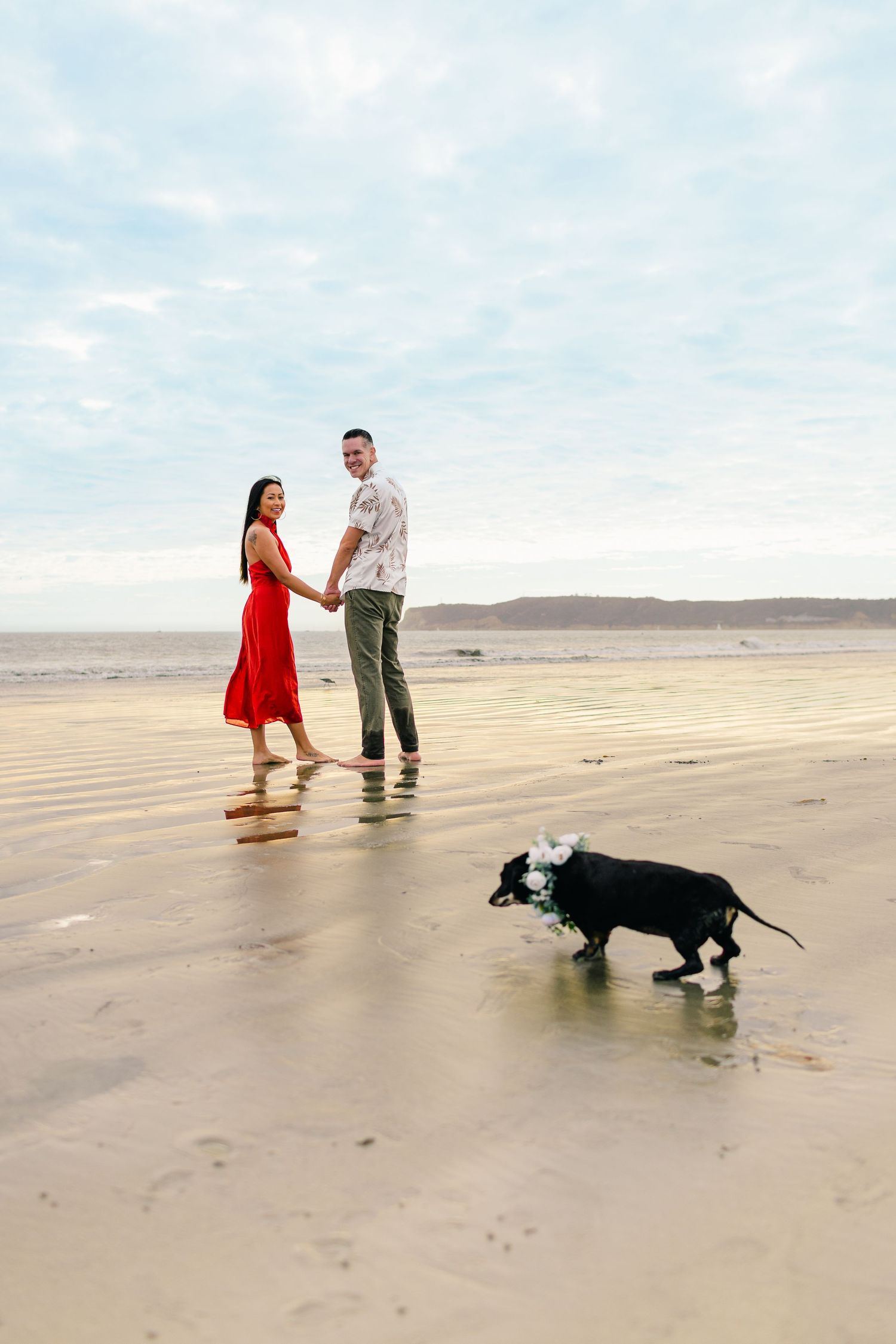 A couple walks along a vast sandy beach with their black dog trailing behind them on a cloudy day.