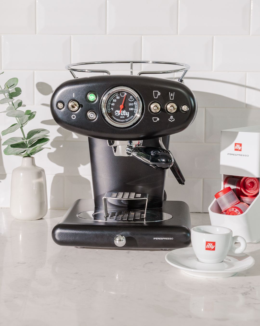 Black illy espresso machine with pressure gauge and cup on white counter with eucalyptus plant.
