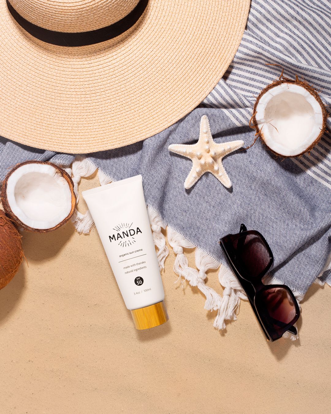 Beach lifestyle product flatlay with sunhat, shells, and Manda skincare products on gray fabric.