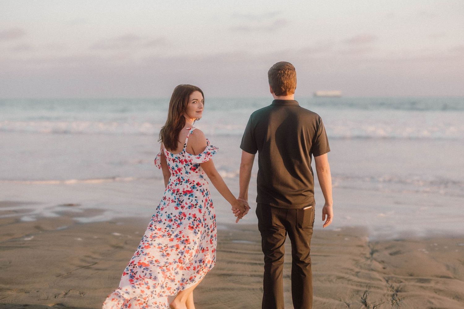 A couple strolls along a sandy beach at sunset with waves crashing in the background.