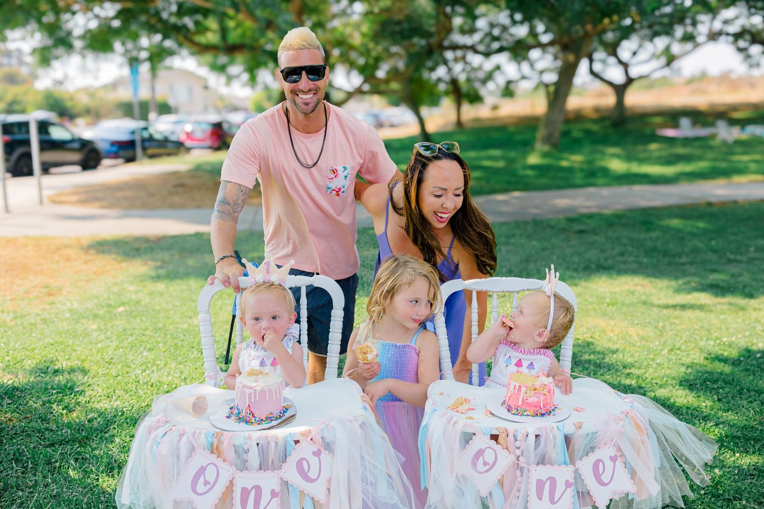 Twins in matching pink tutus celebrate their first birthday party outdoors with family in a park setting.