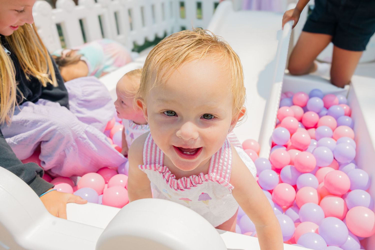 Baby playing in a ball pit filled with pink and purple plastic balls at an outdoor party.