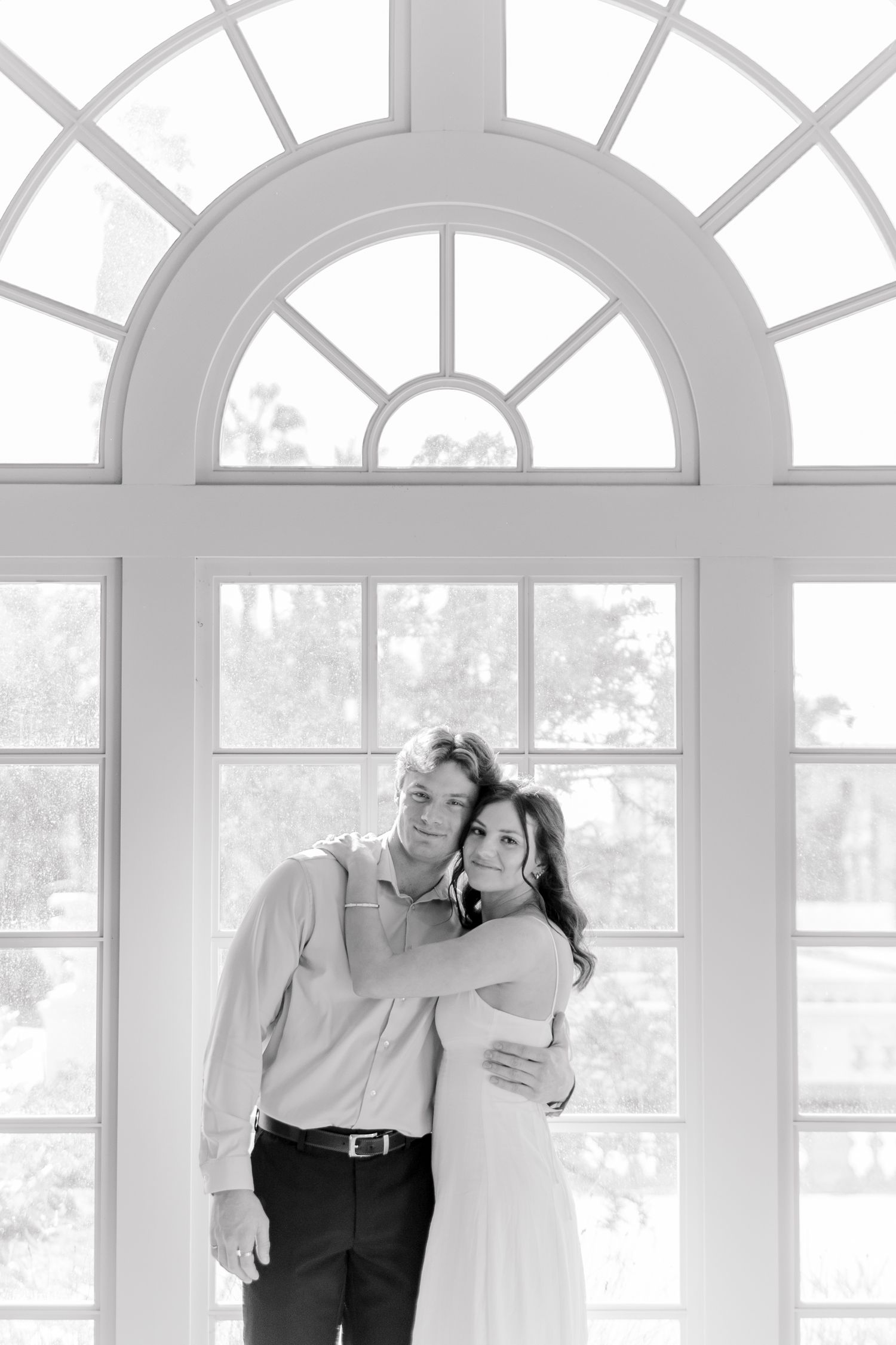 A romantic black and white portrait in front of a large arched window with geometric panes.