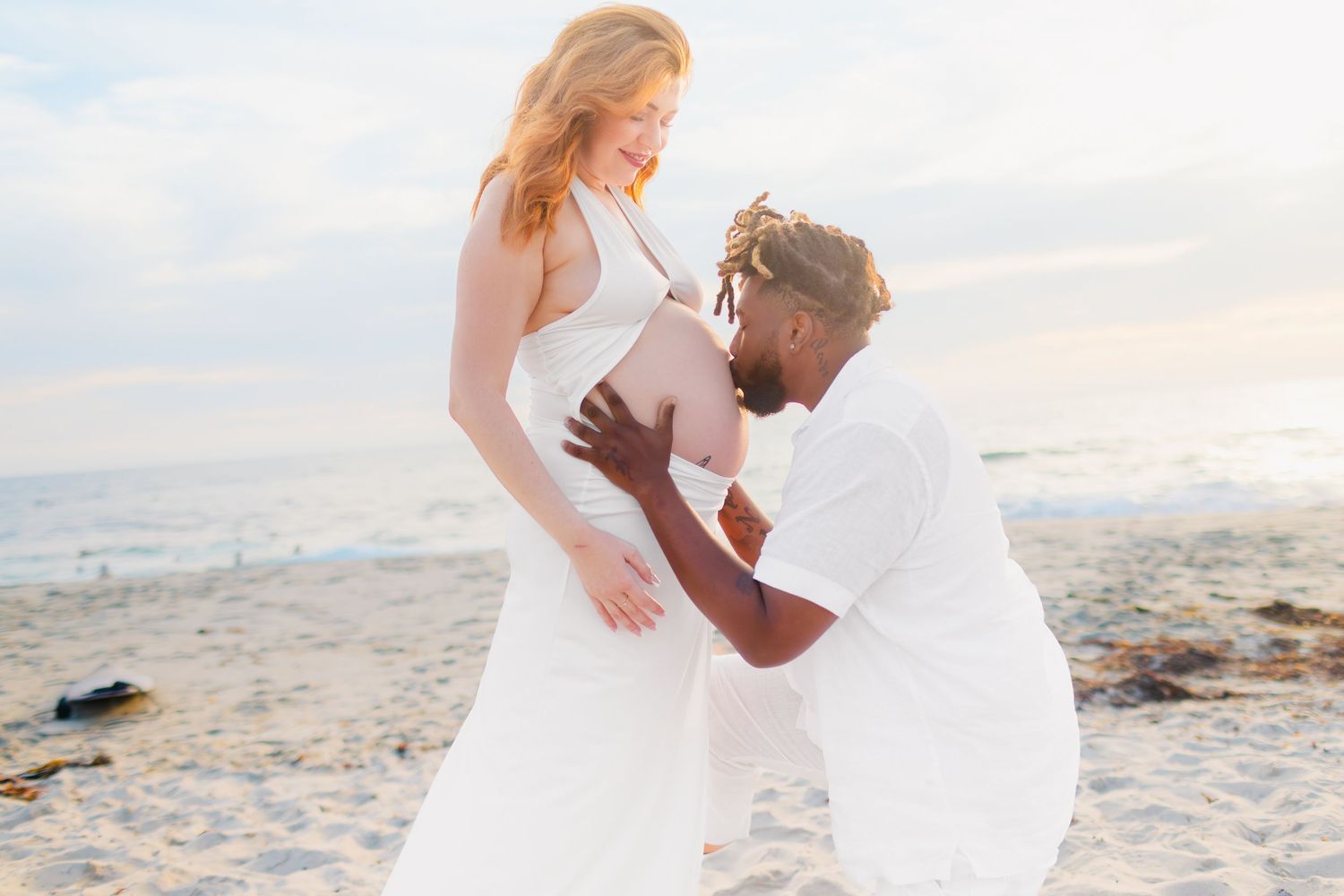 A couple embraces on a sandy beach at sunset while wearing white wedding attire during a romantic photoshoot.