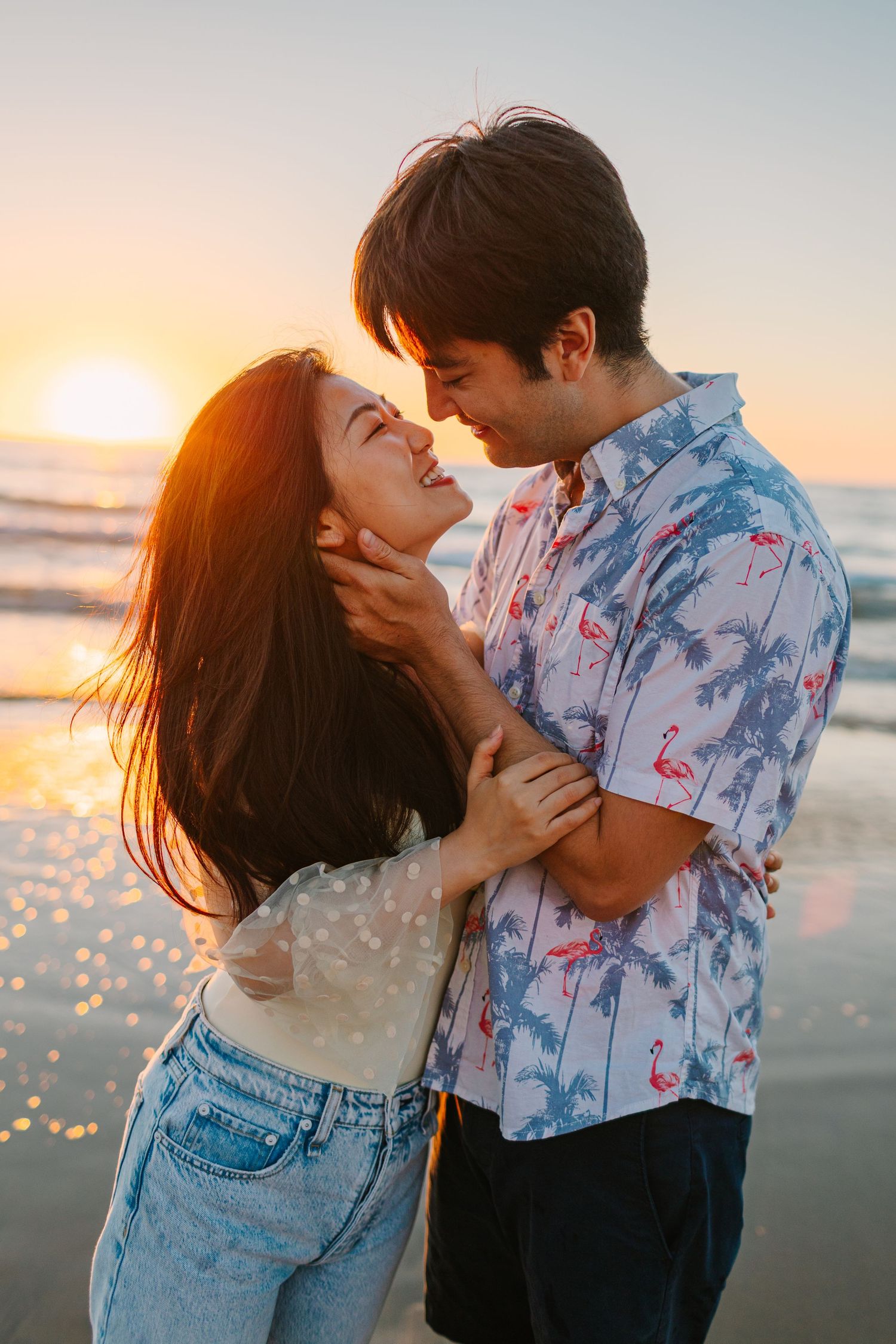 Couple shares tender moment on beach at sunset with waves and golden light in background.