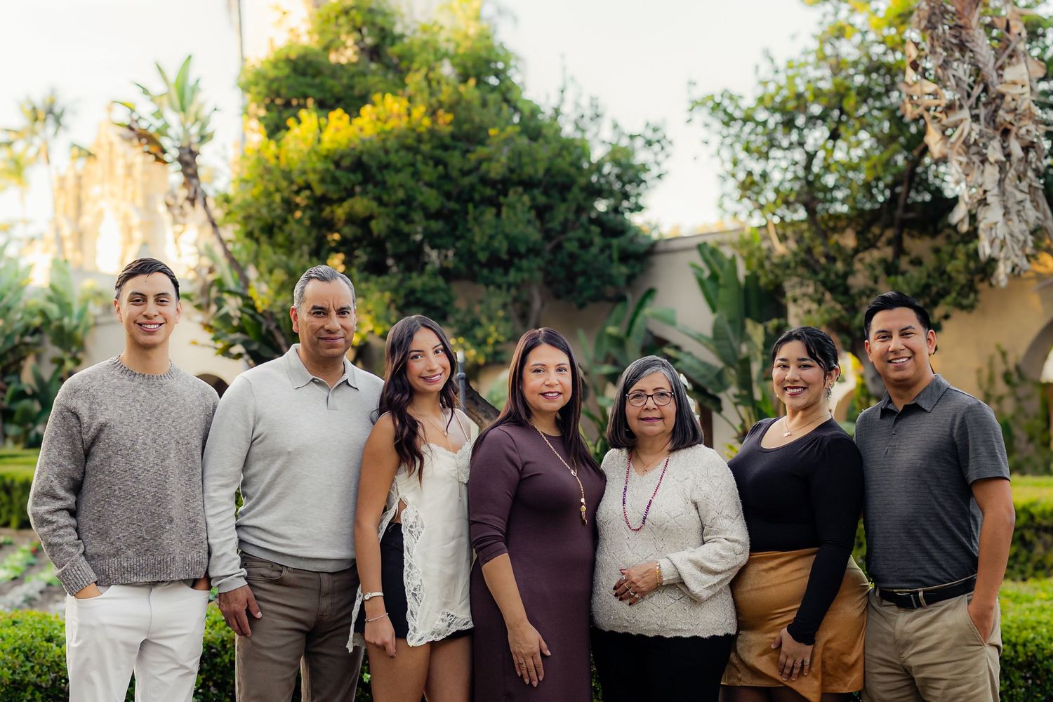 A family group stands together outdoors in coordinated grey and white outfits against a natural backdrop.