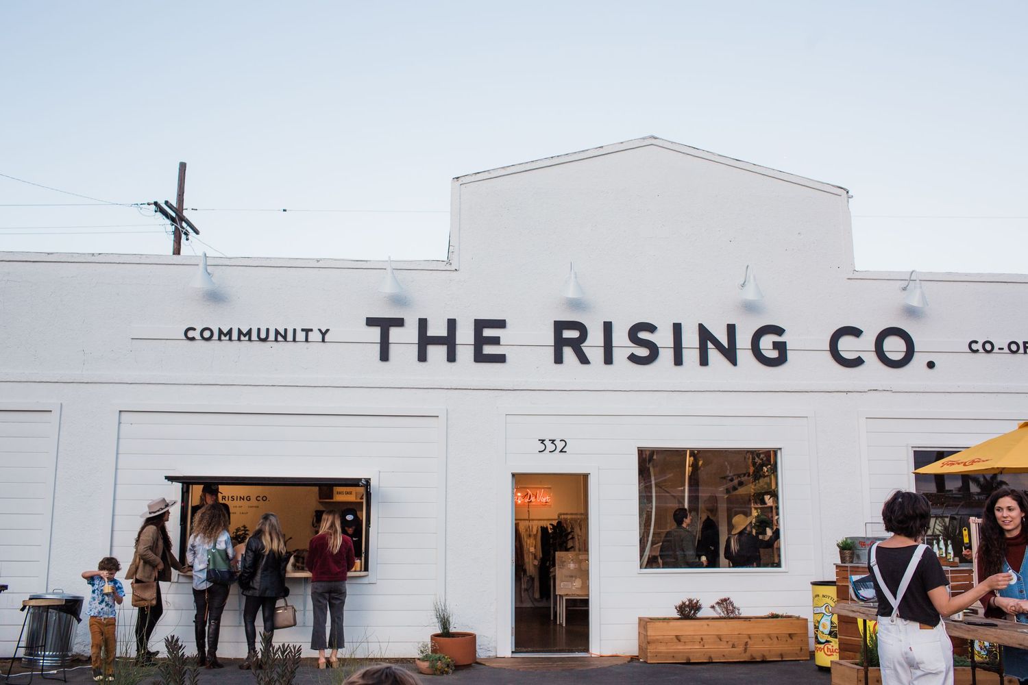 White industrial storefront of The Rising Co. community space with customers visible through large windows at sunset.