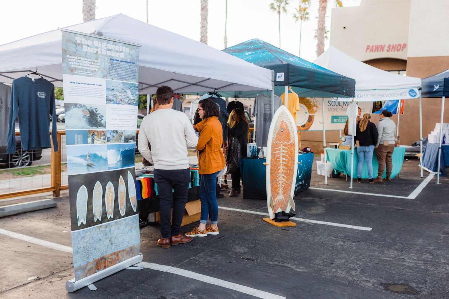 Visitors walking through an outdoor marketplace with white tents and vendor booths set up along a street on a cloudy day.