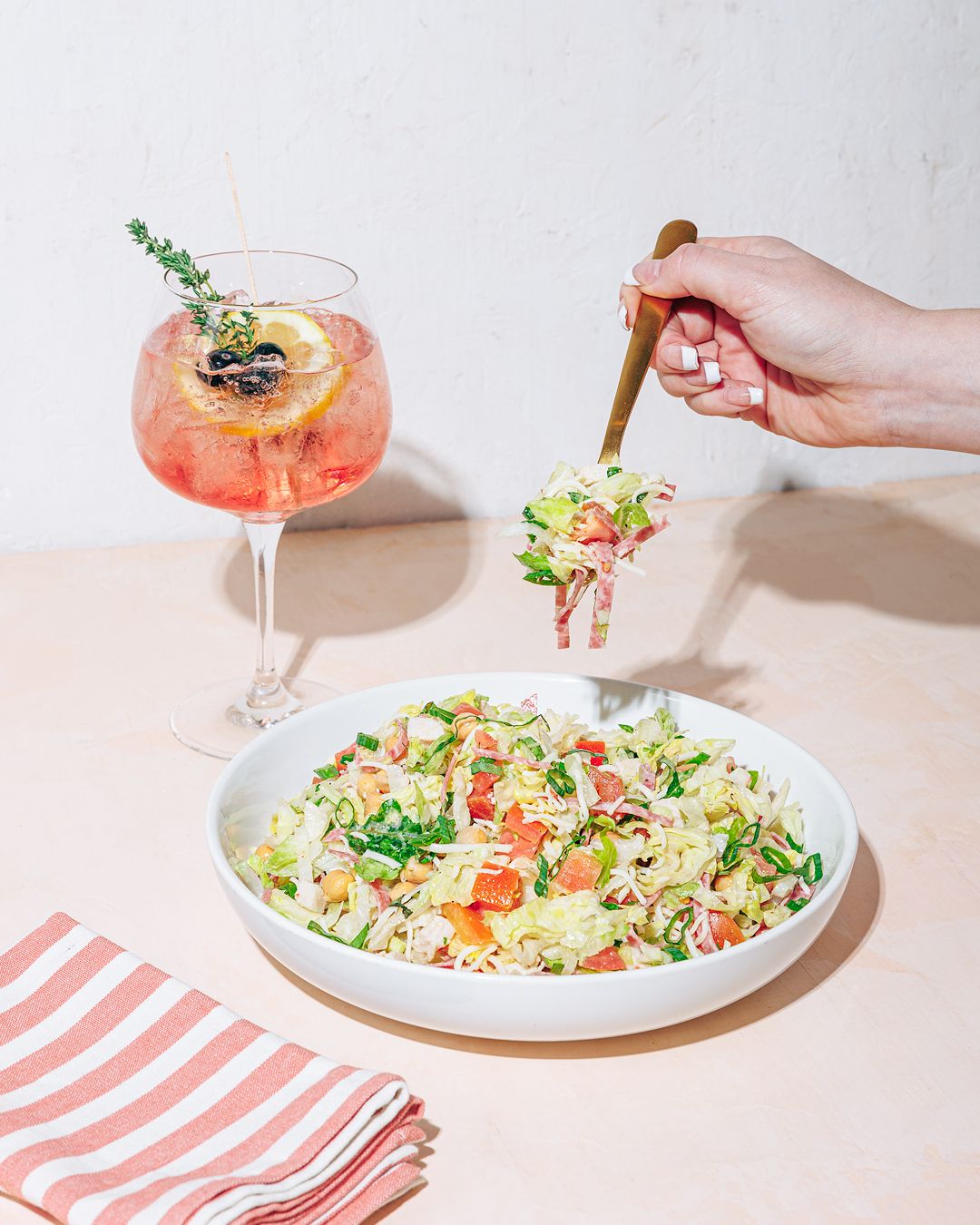 Hands serving colorful salad plates and cocktails on a pink and white striped table setting.
