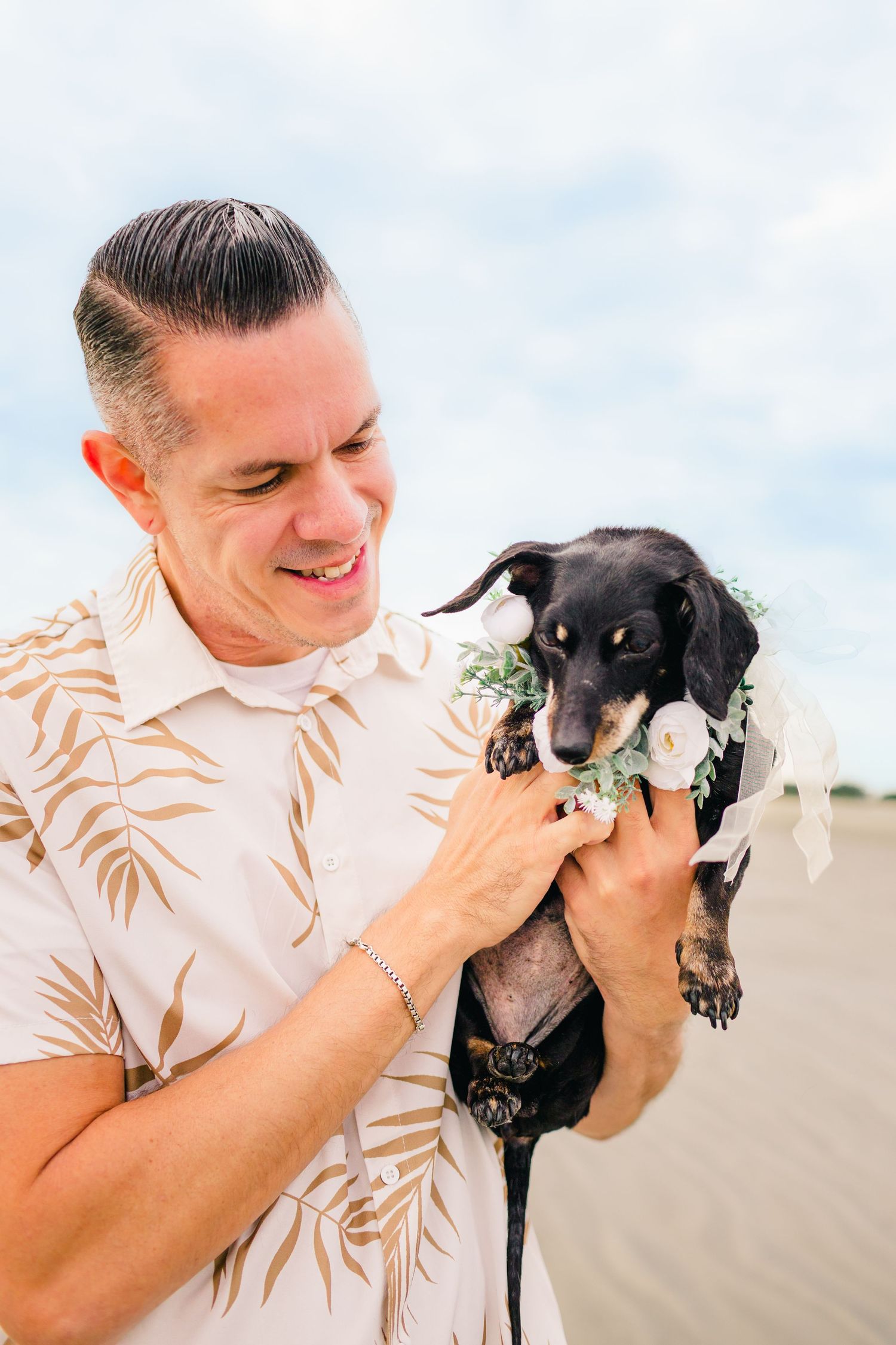 Sweet moments captured as someone holds and cuddles a small black dog wearing a floral collar on a sunny beach day.