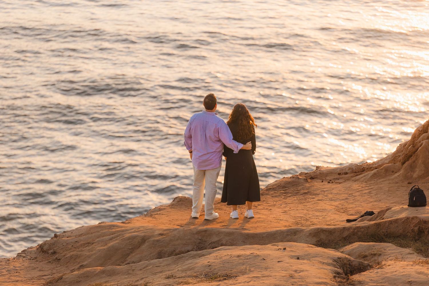 Two people pose together on an oceanside cliff at sunset with waves crashing below.