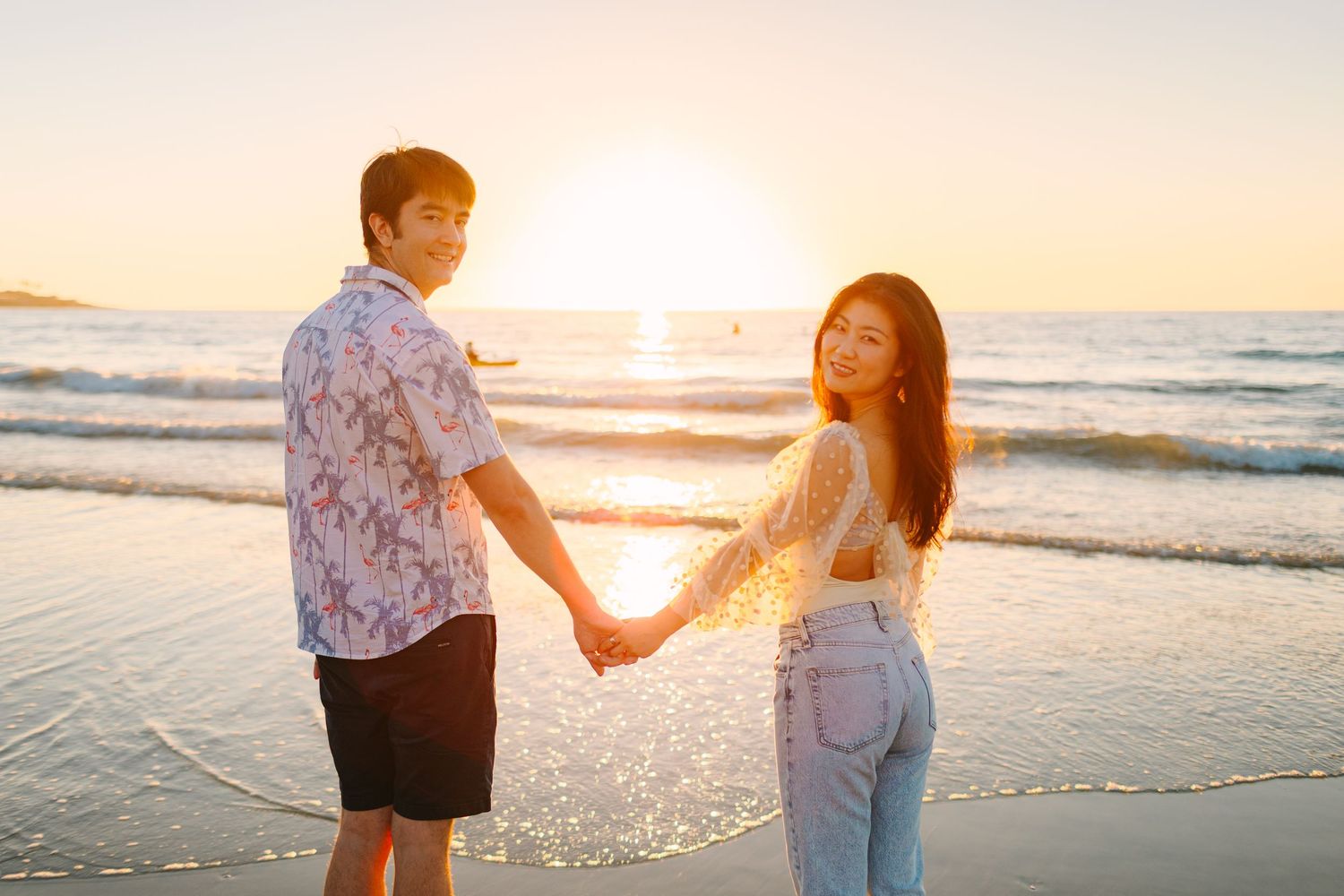 A couple walks along a sunset beach holding hands as golden light illuminates the ocean waves.