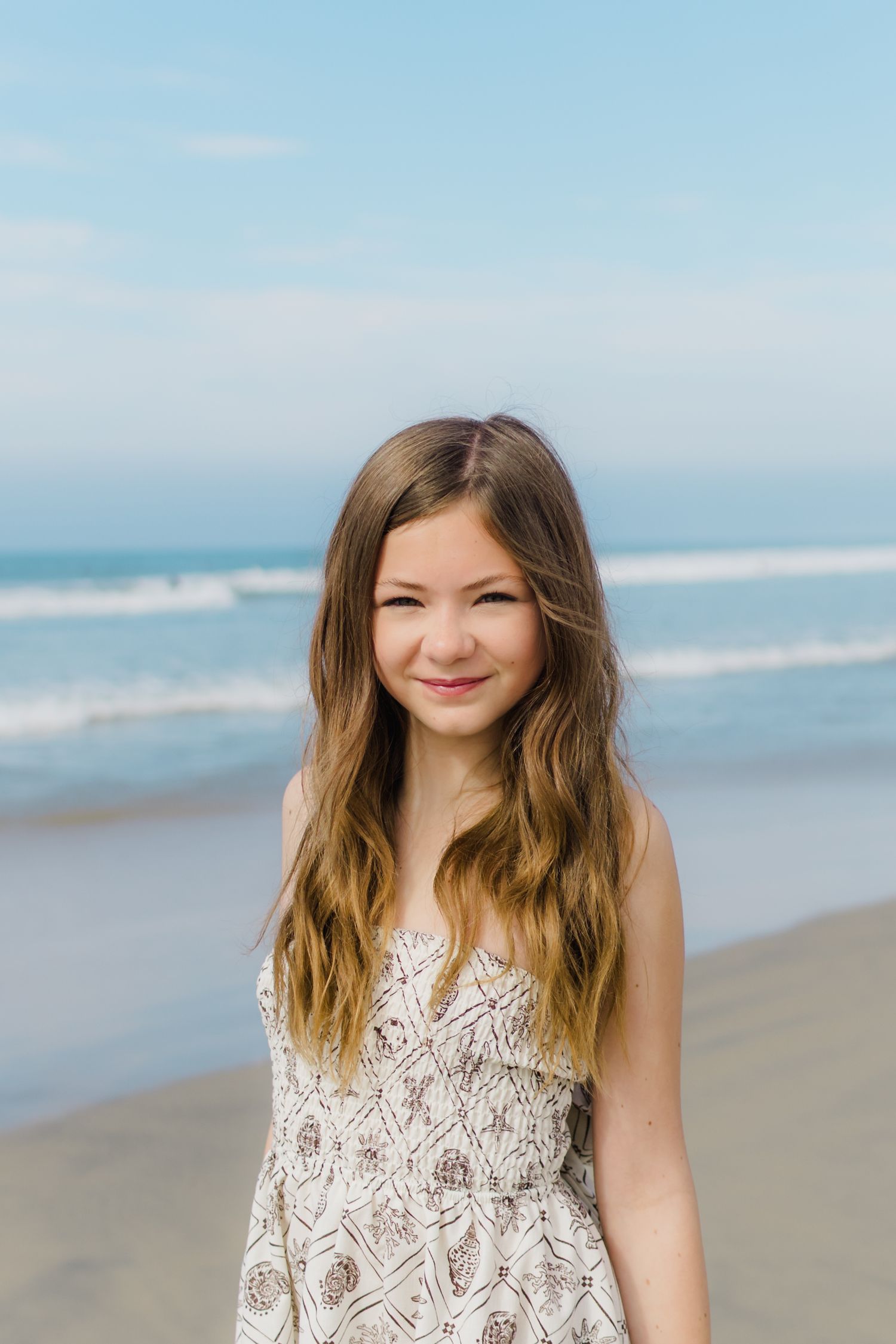Young woman in white lace dress smiling on sandy beach with ocean waves and blue sky.