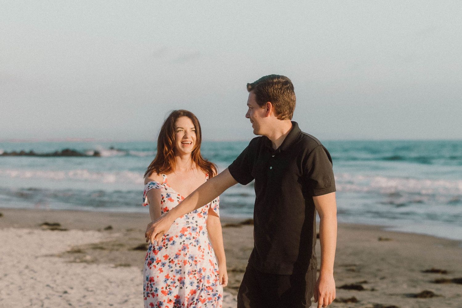 Happy couple laughing and holding hands while strolling on beach near luxury hotel.
