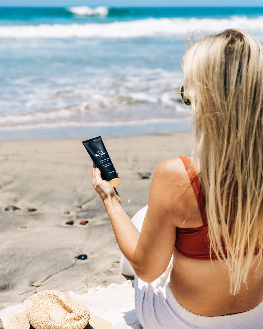 Person holding black Manda skincare tube while relaxing on sandy beach by ocean.