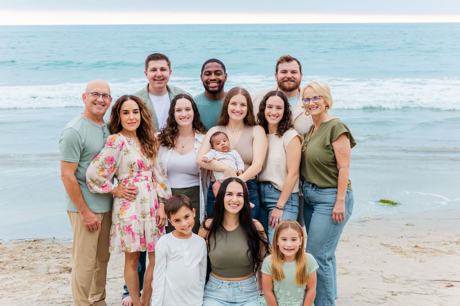 A large extended family gathers for a group photo on the beach with waves in the background.
