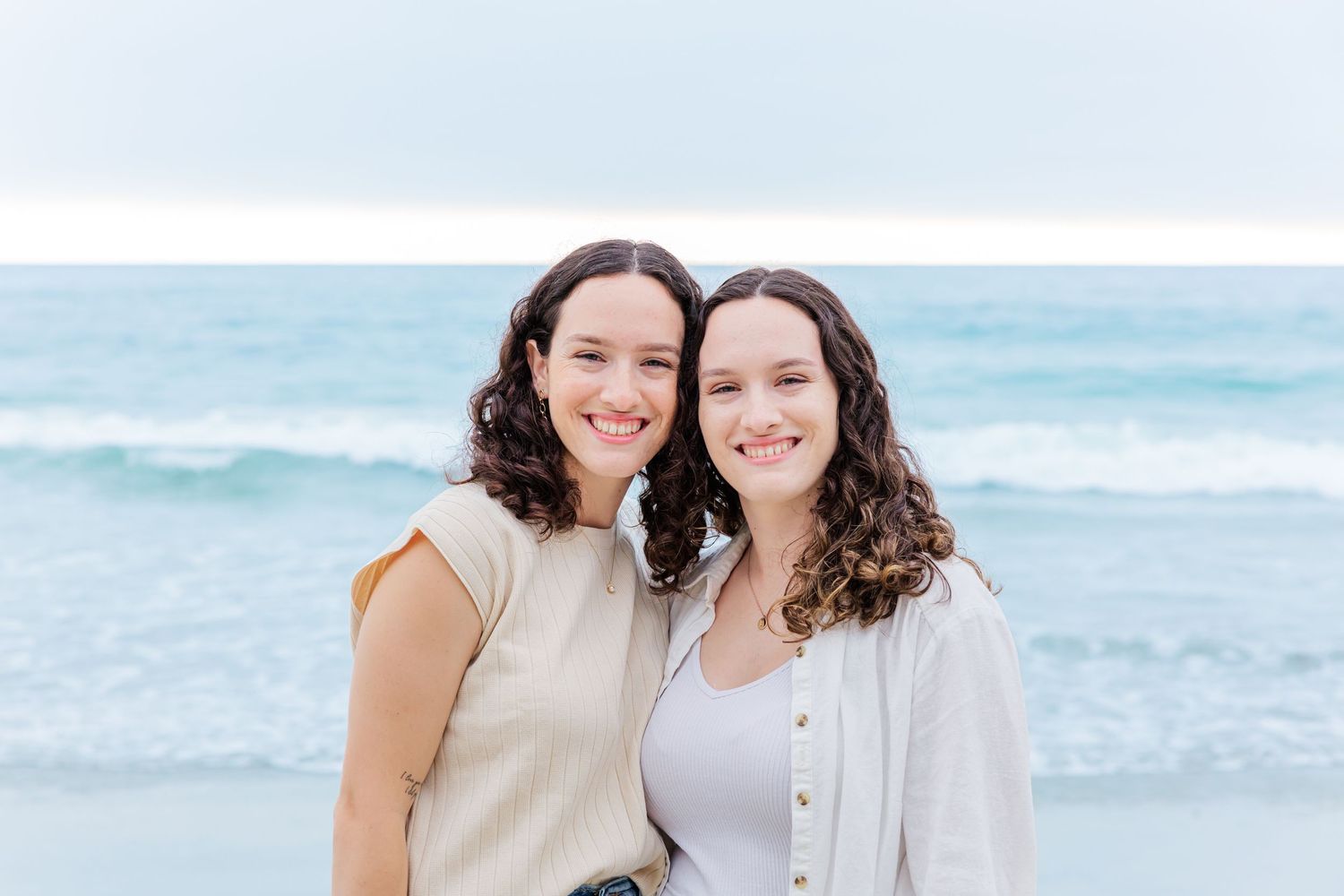 Two friends share a warm moment together on the beach with ocean waves in the background.