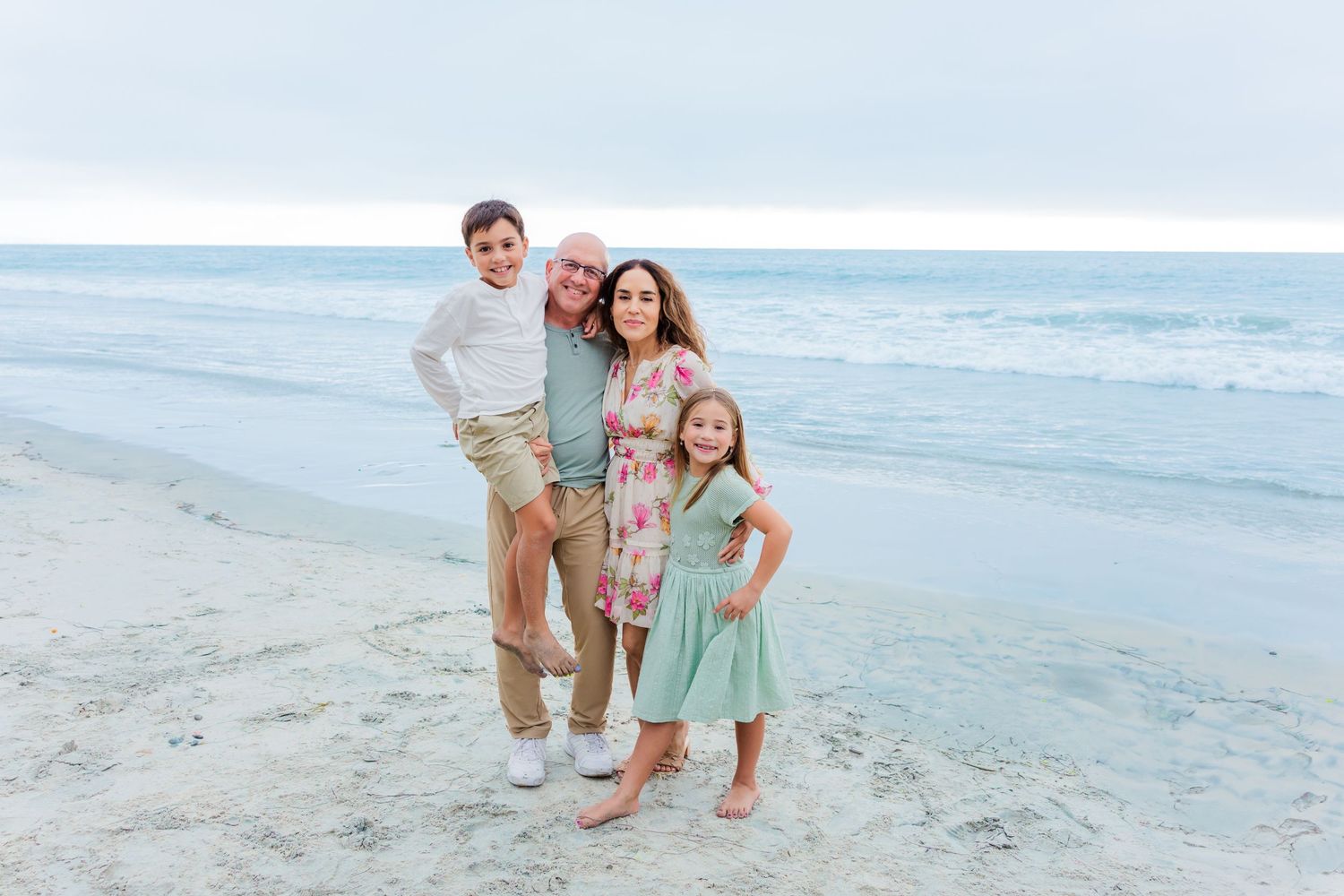 A family of four poses together on a sandy beach with ocean waves in the background.