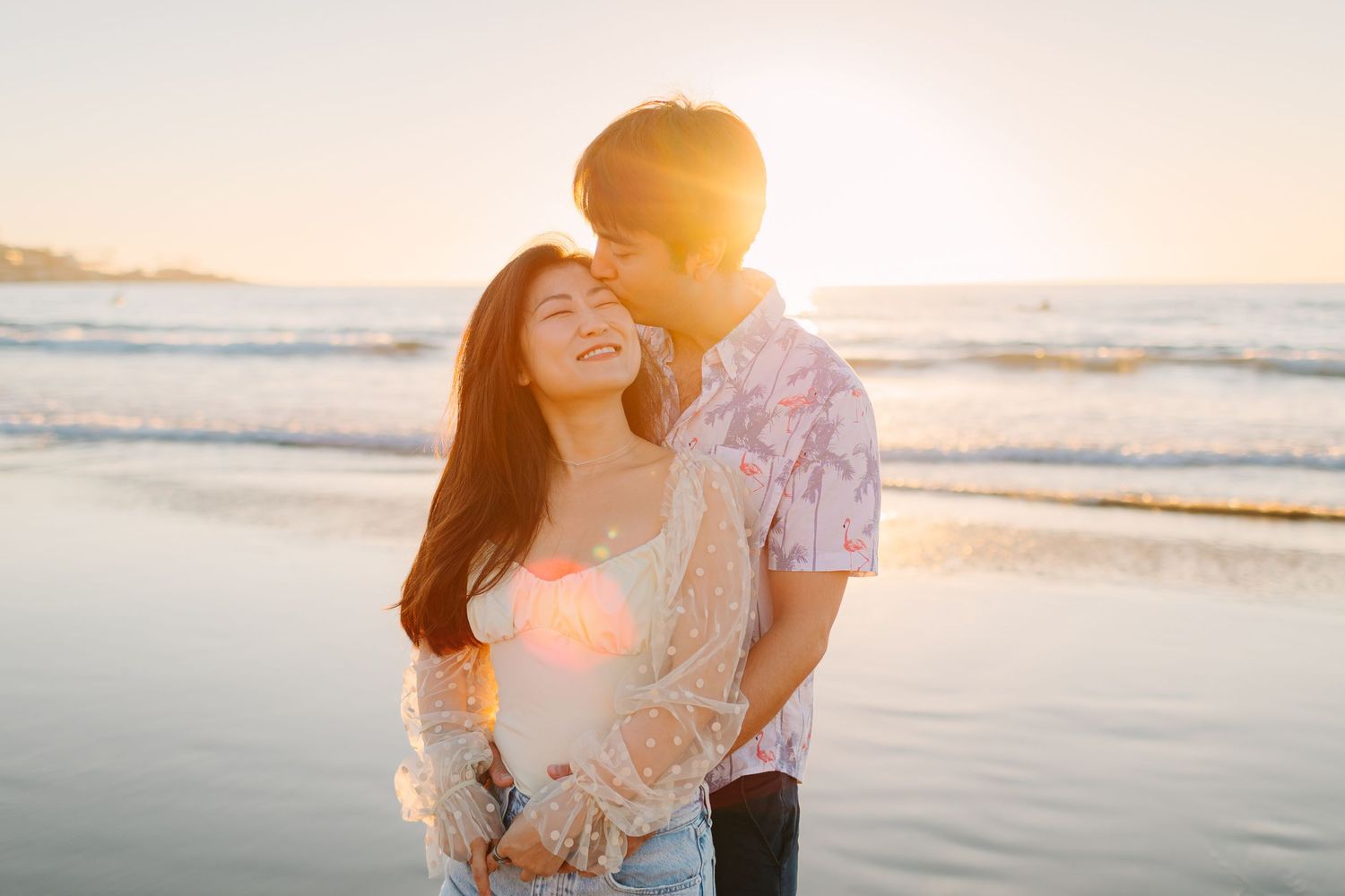 A couple embraces on the beach during golden hour as waves roll onto the shore in the background.