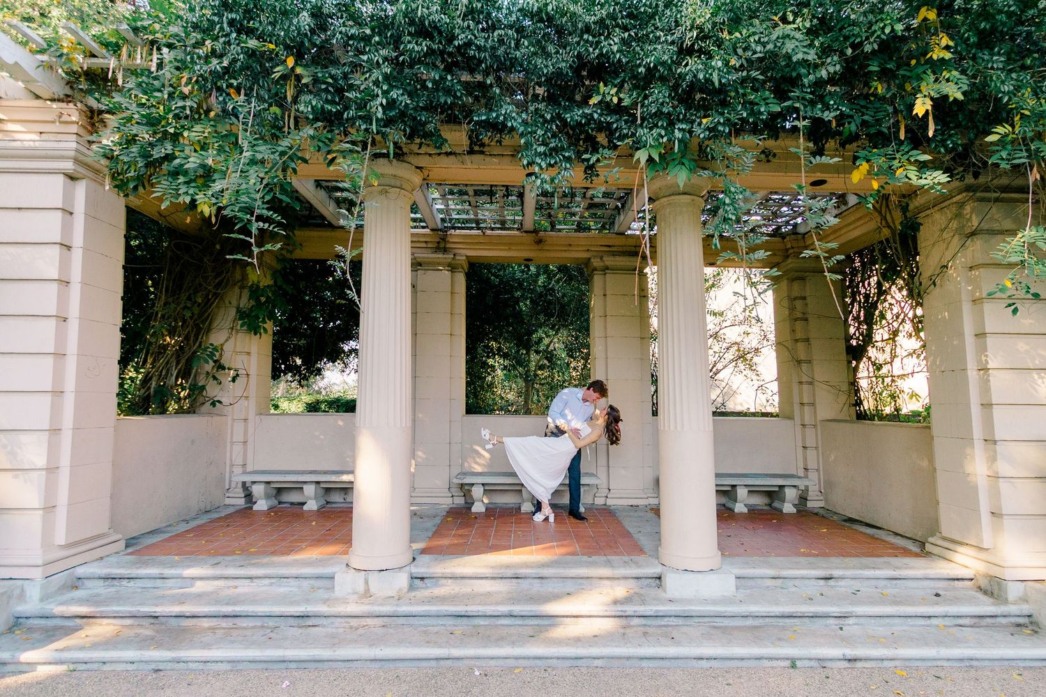 A long covered walkway with white columns and green vines growing overhead creates a shaded path through a garden.