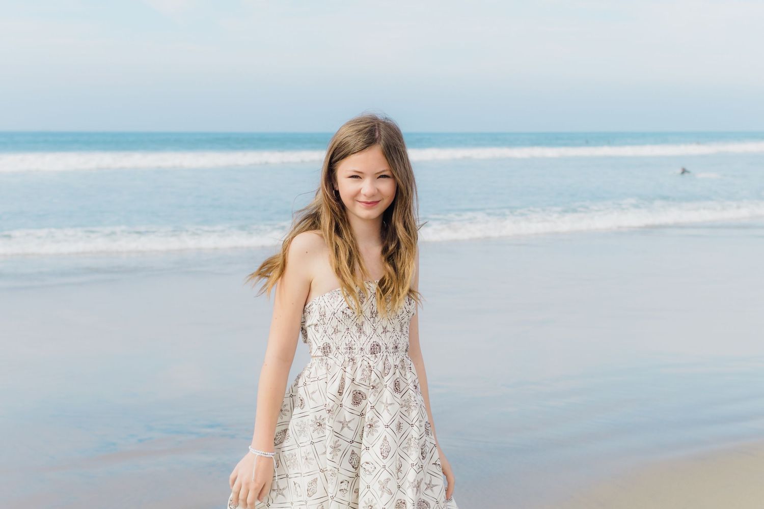 A person in a white lace dress walks along a peaceful beach with soft waves and cloudy sky in the background.