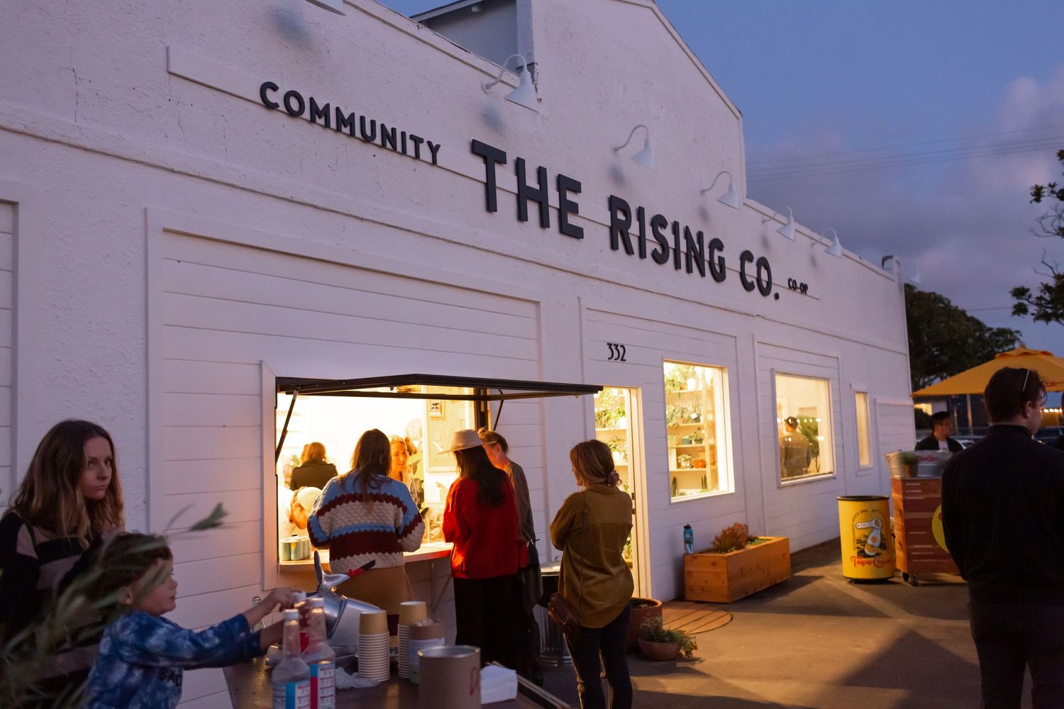 People gather outside lit storefront windows of The Rising Co during evening hours in Oceanside, California.