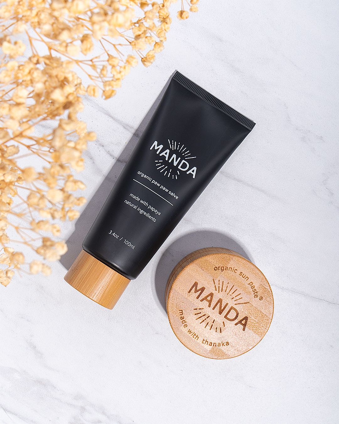 Black Manda skincare tubes and wooden jar lids arranged on white marble surface.
