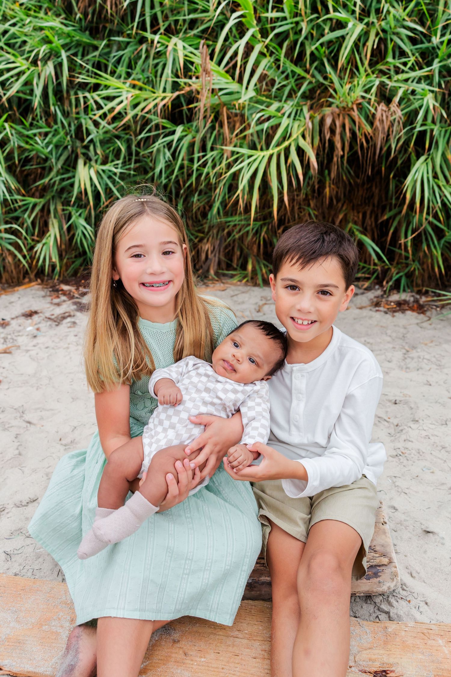 Three siblings sit together against a bamboo backdrop, with a small baby in their arms.