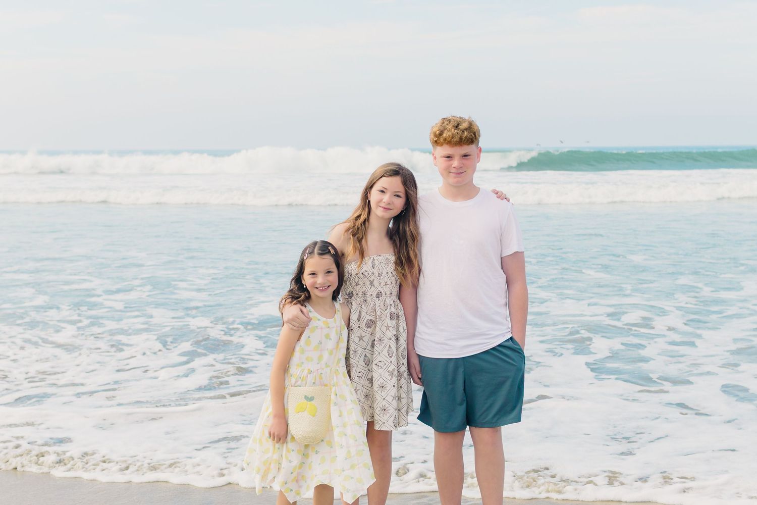 Three people stand together smiling on a sunny beach with ocean waves rolling in behind them.