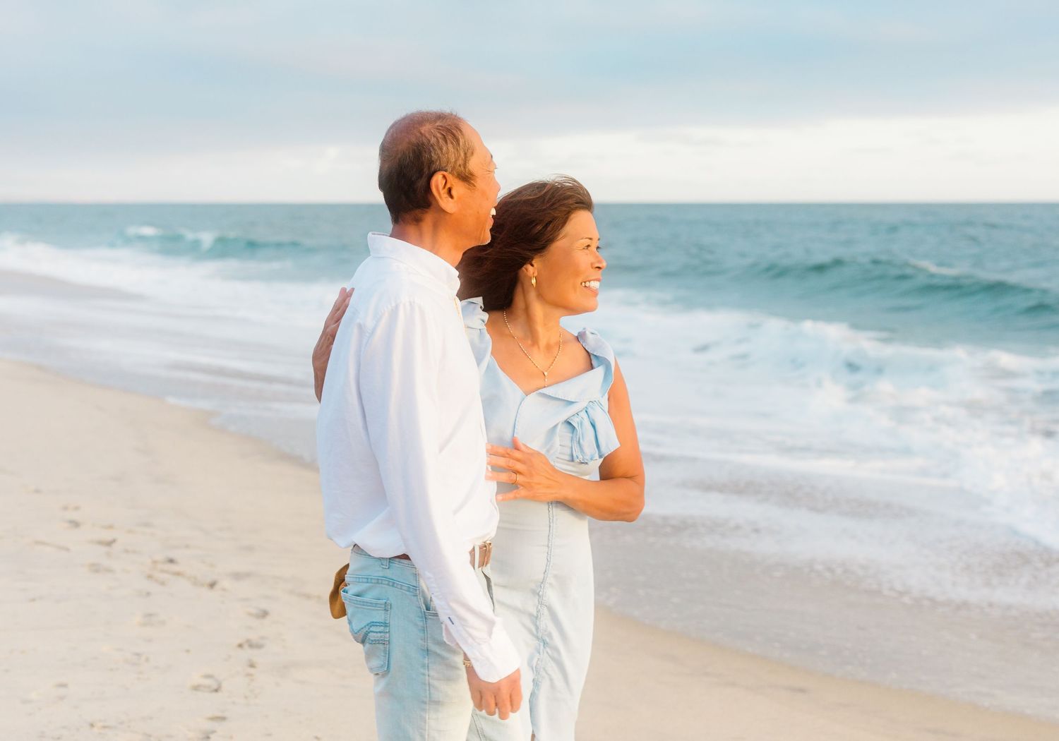 A couple walks along a scenic beach at sunset wearing white and light blue attire.