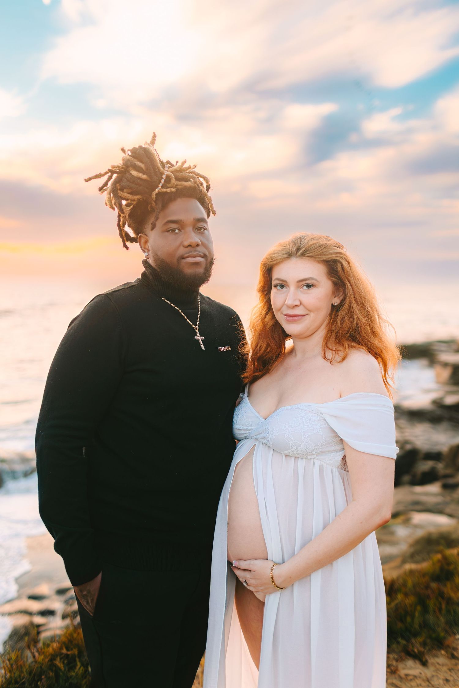 A couple shares an intimate moment during a sunset photoshoot on a coastal cliff with soft pastel skies in the background.