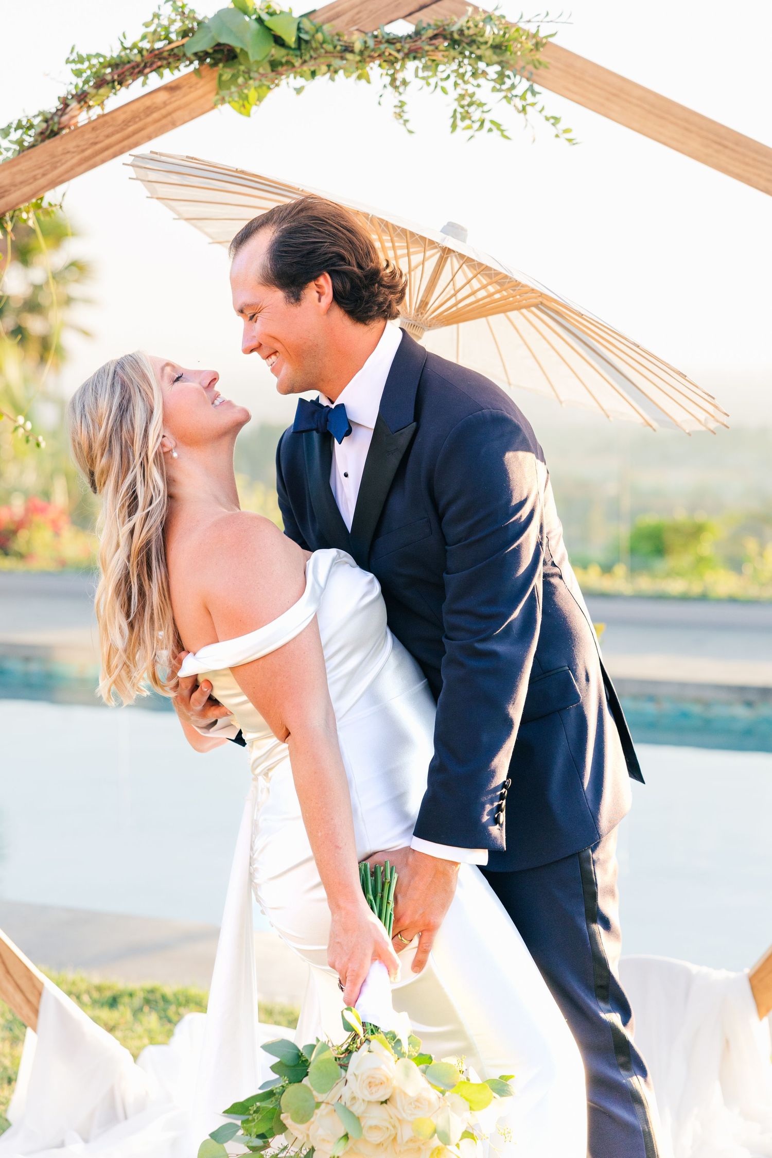 Wedding couple shares romantic kiss under floral arch with waterfront view.
