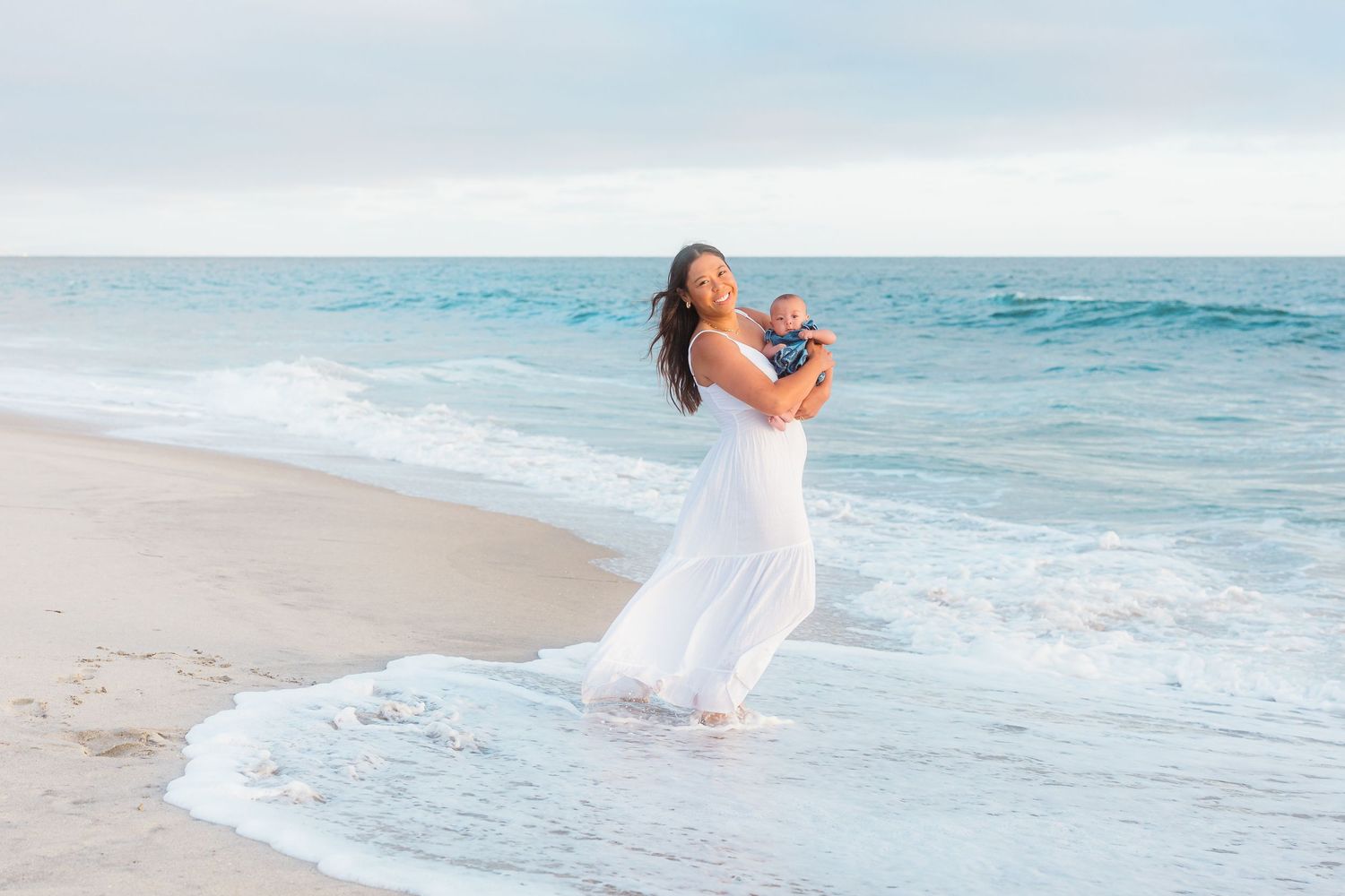 A woman in white holds a baby up joyfully at the beach with waves rolling in.