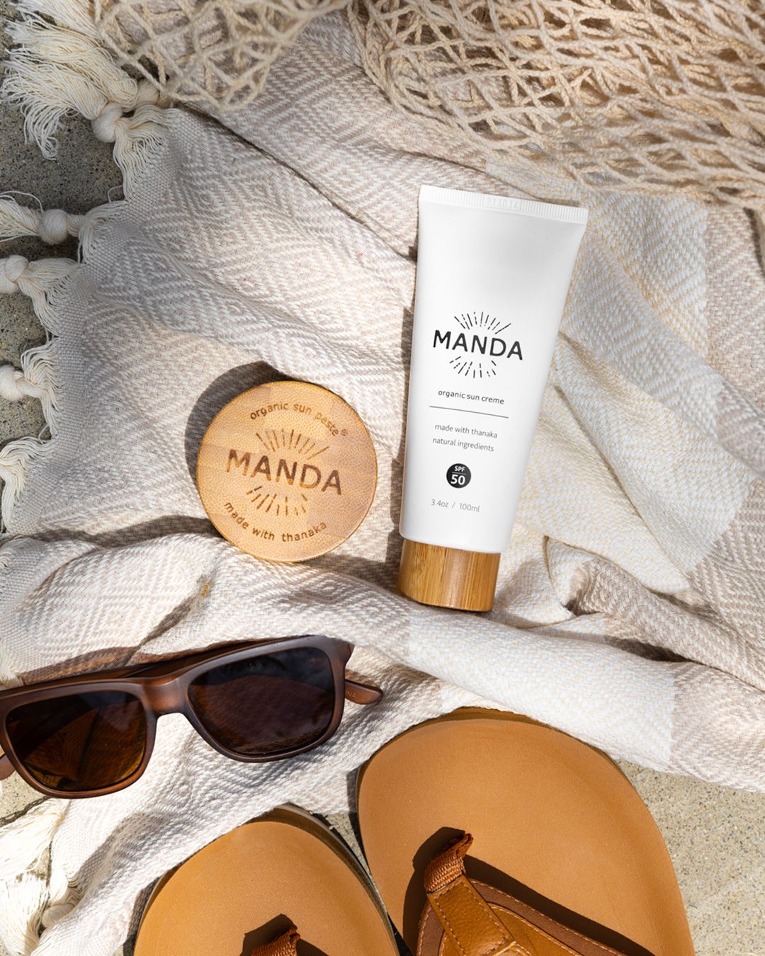 Manda skincare products arranged with sunglasses and sandals on textured white fabric.