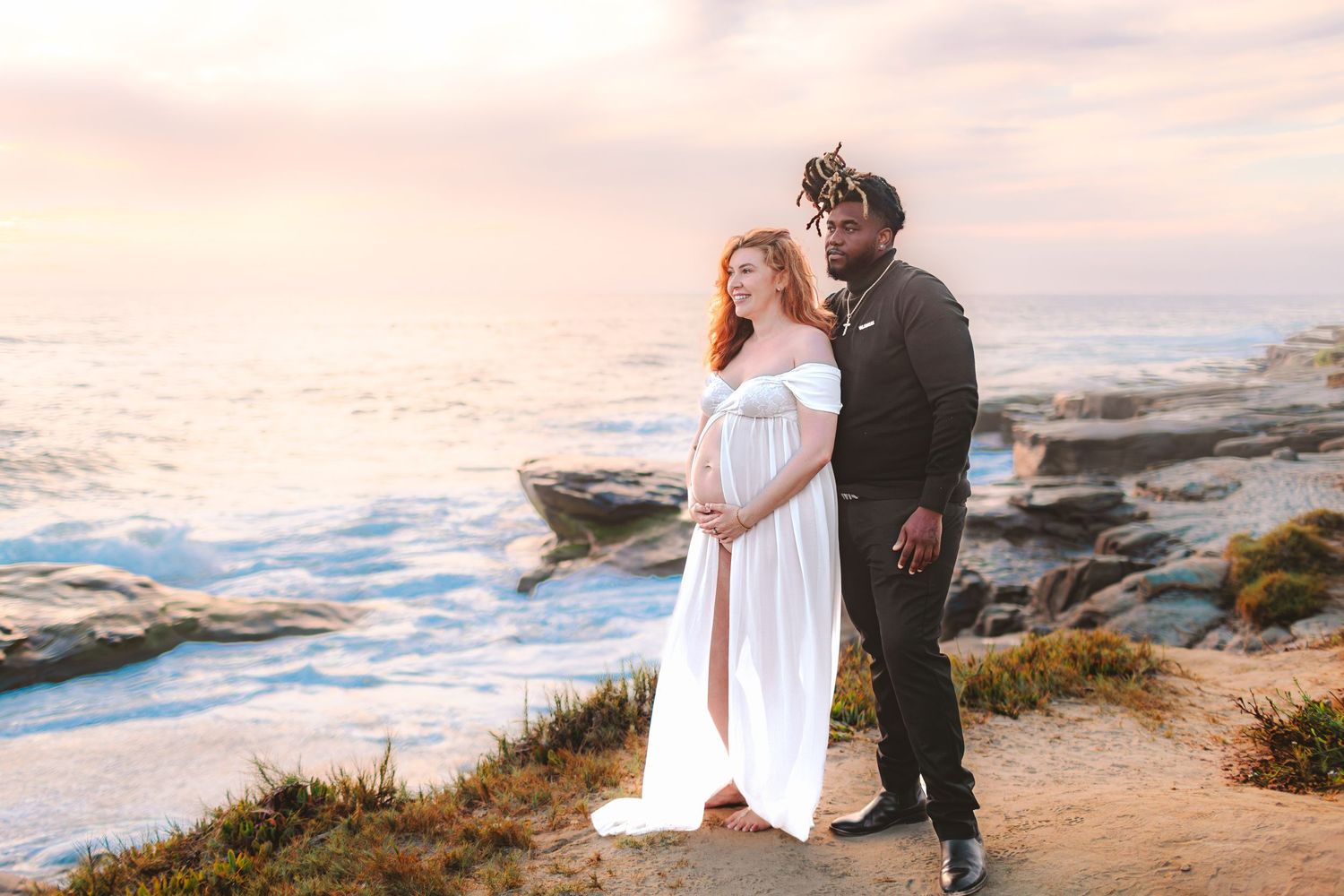 A couple stands on a rocky coastal cliff at sunset, with the woman wearing a flowing white dress and man in dark attire.