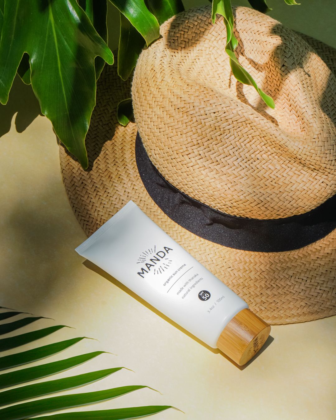 White Manda skincare tube displayed with straw hat and tropical palm leaves.
