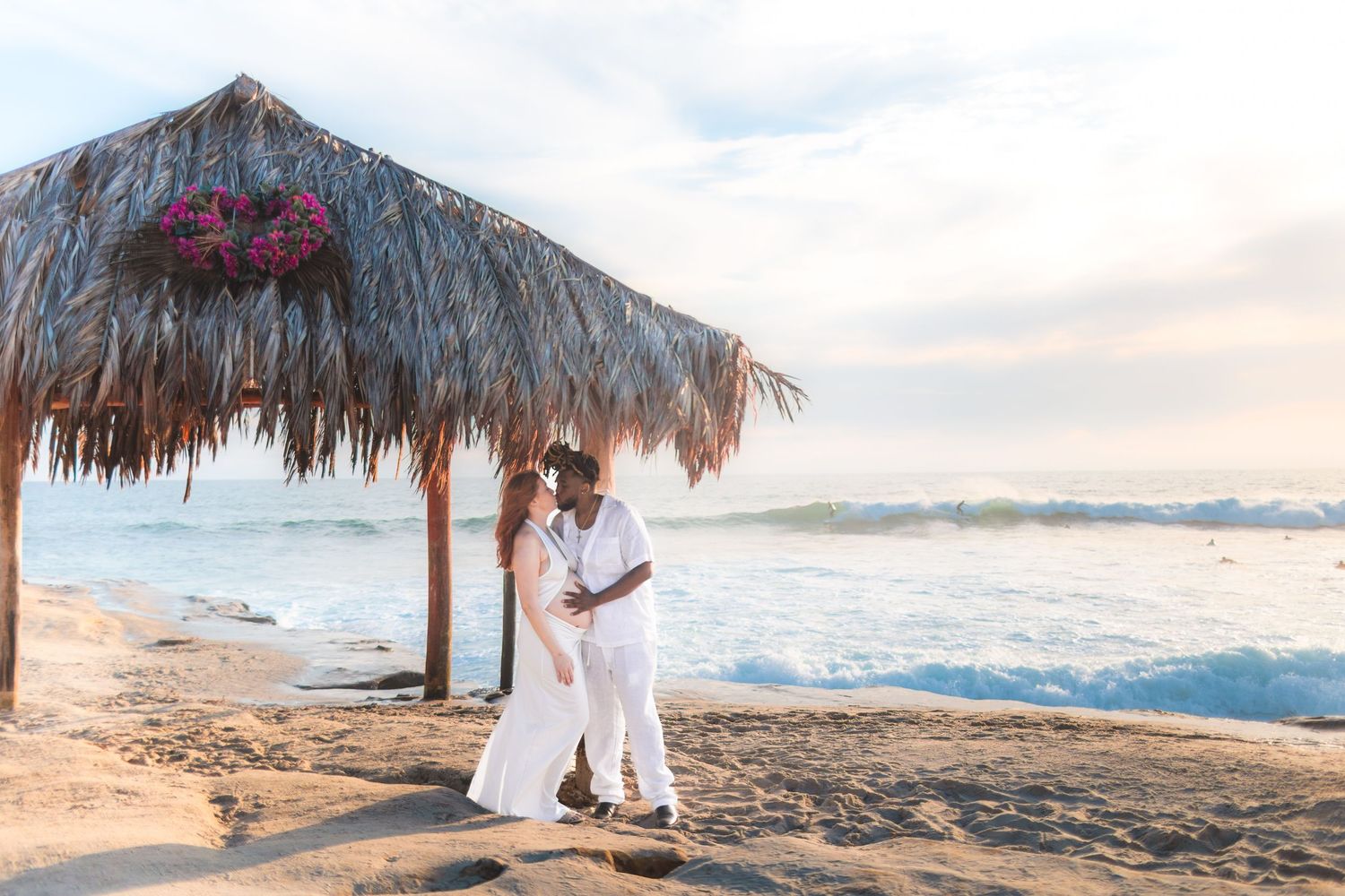 A romantic couple in white outfits embraces under a thatched beach palapa as ocean waves roll in at sunset.