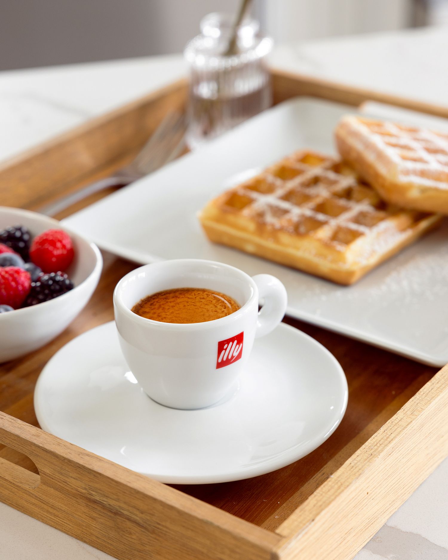 Wooden breakfast tray with illy espresso coffee, fresh berries, and golden waffles served on white plates.