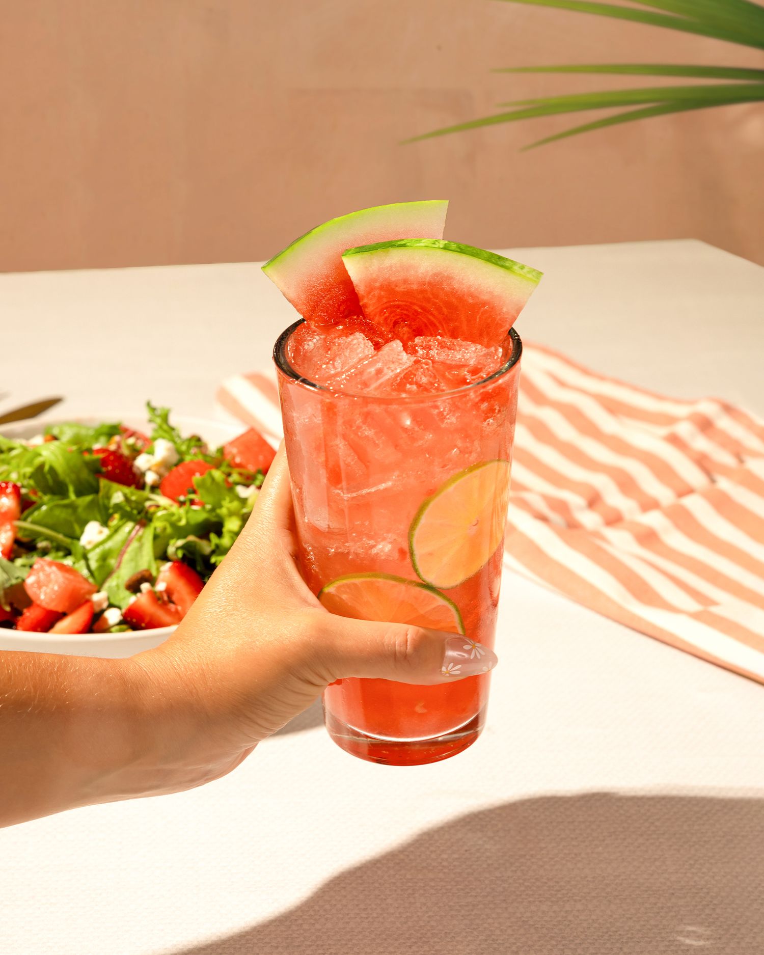 Pink cocktails and fresh salads served on a striped tablecloth in natural light.